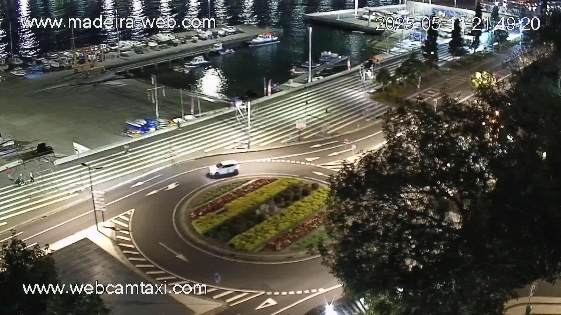 A nighttime aerial view shows a car navigating a roundabout near a harbor with boats docked, under a clear sky.