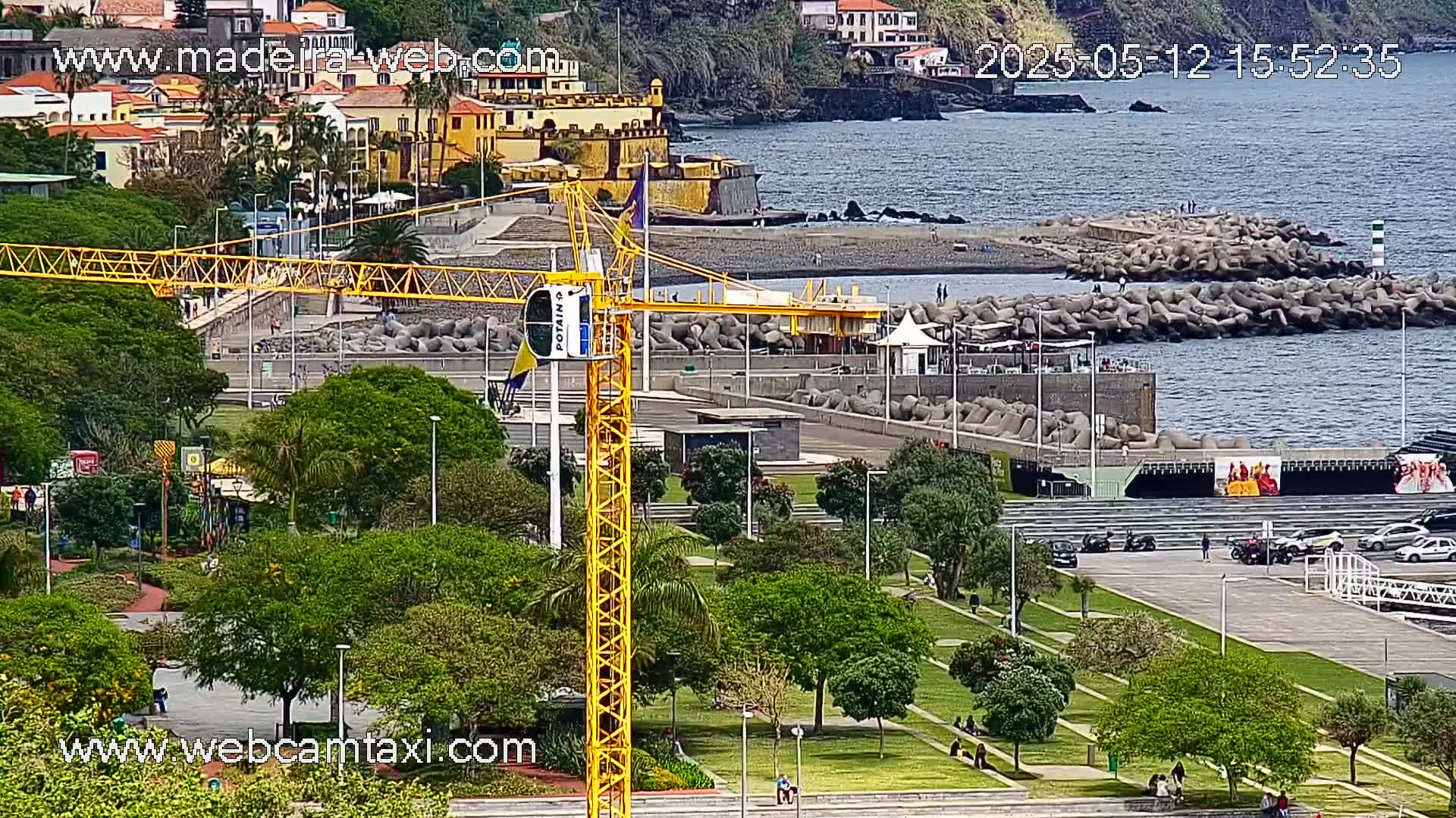 A yellow crane stands in a park overlooking a coastline with a rocky breakwater and buildings on a hillside under partly sunny skies.