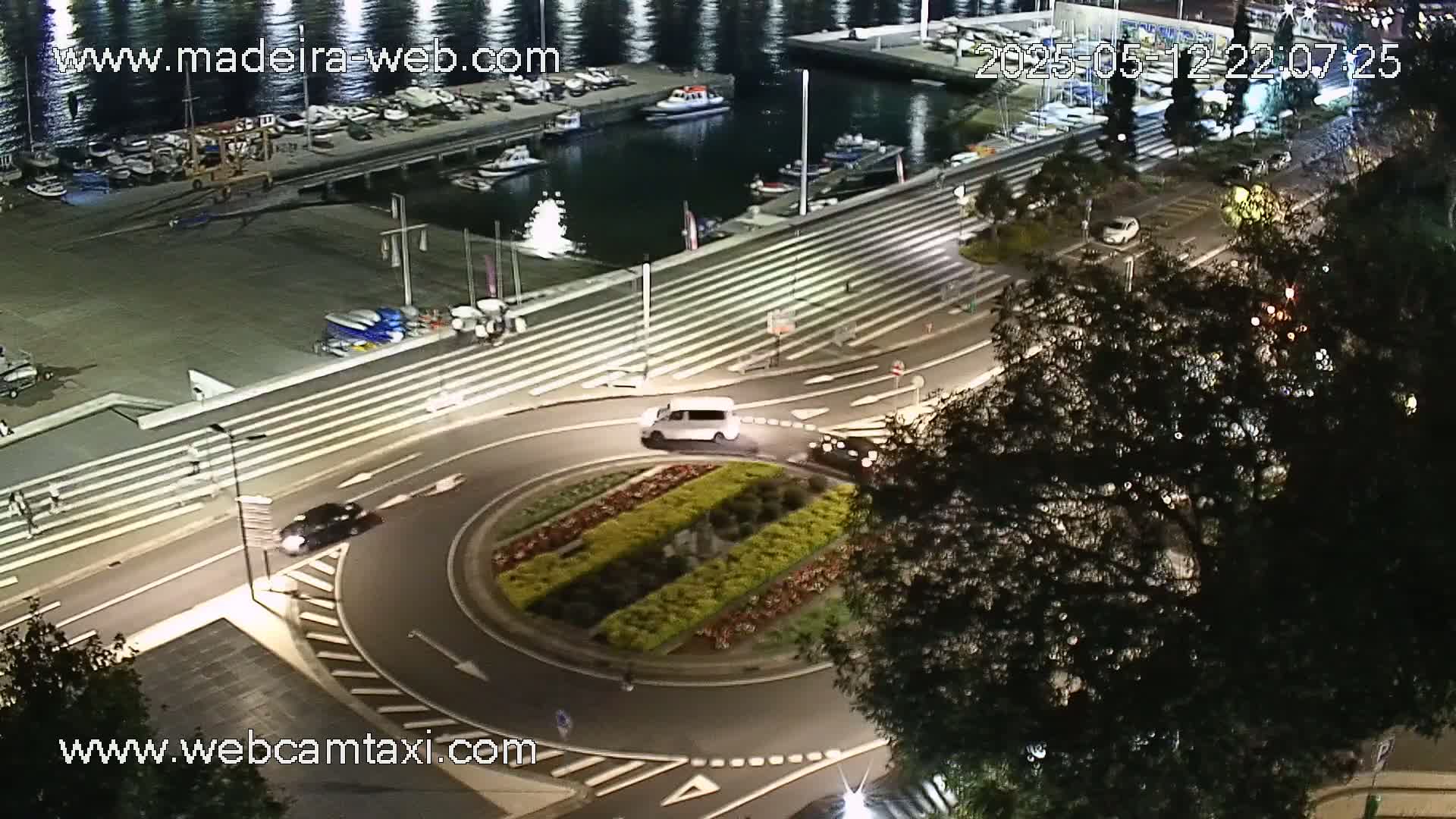 An aerial nighttime view shows a roundabout with a landscaped center, several cars traveling on roads surrounding a harbor with docked boats.