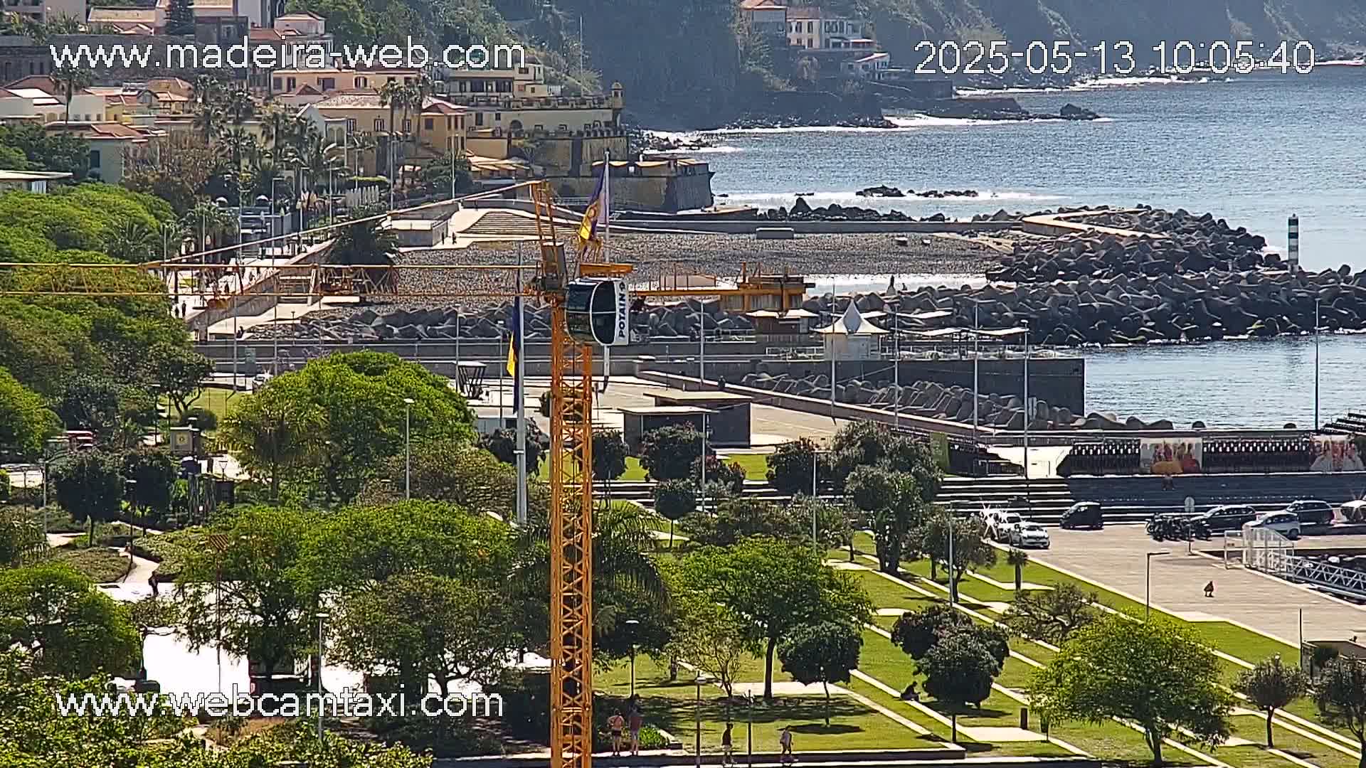 A coastal cityscape on a sunny day features a park with a construction crane, a pebble beach protected by a breakwater, and buildings along a hillside overlooking the ocean.