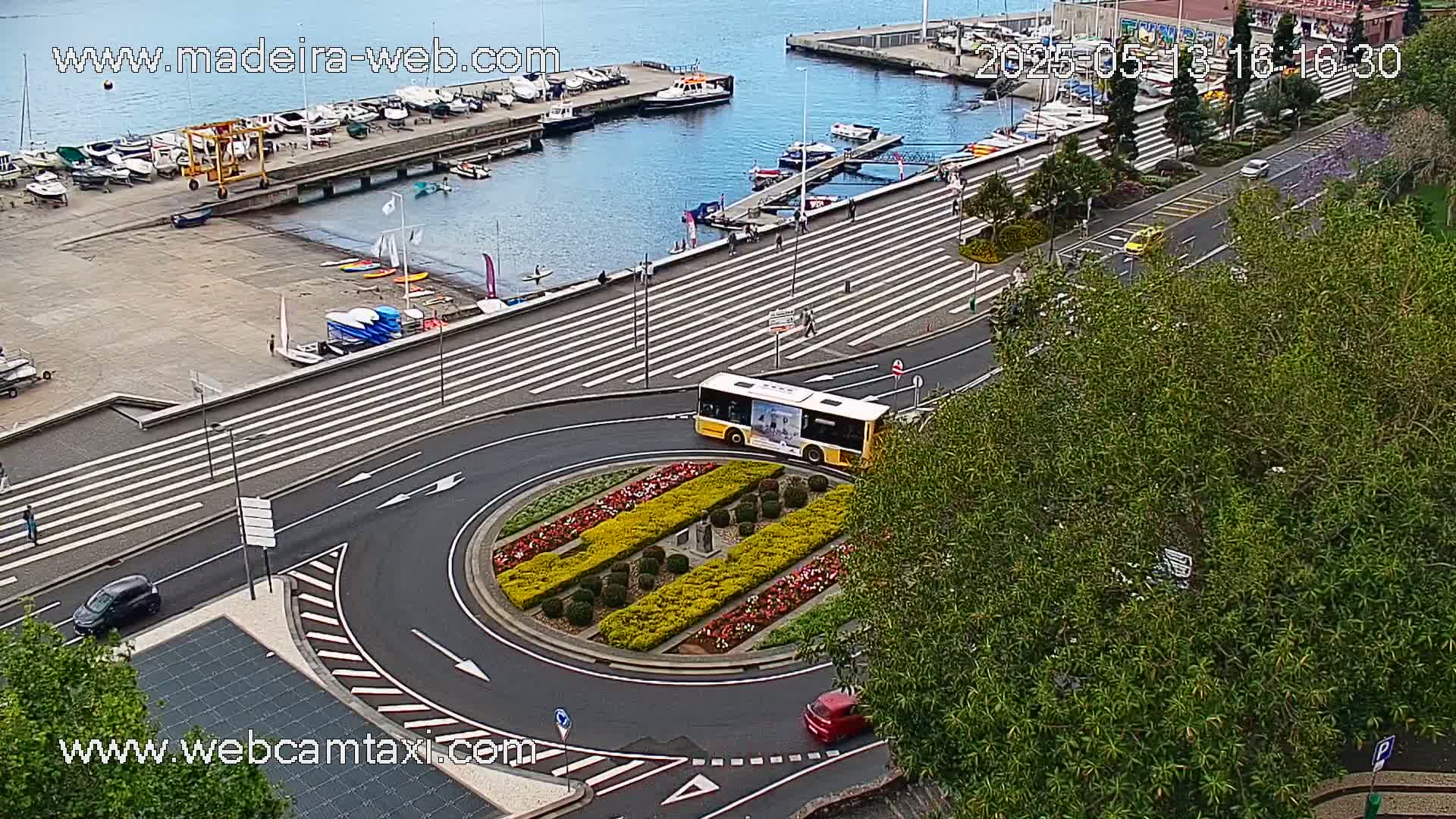 A yellow bus circles a roundabout decorated with colorful flowers, near a harbor filled with boats on a partly cloudy day.