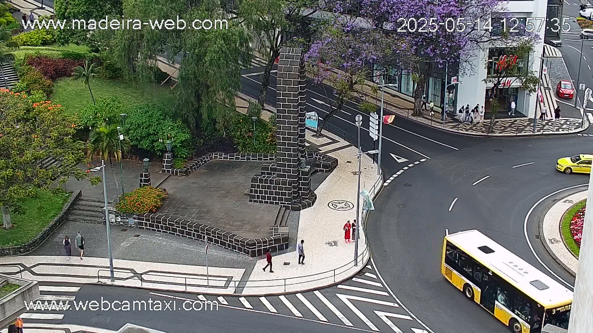 A high-angle view shows a city square with a dark stone monument at its center, surrounded by lush greenery, pedestrians, and a roadway where a yellow bus is turning a corner on an overcast day.