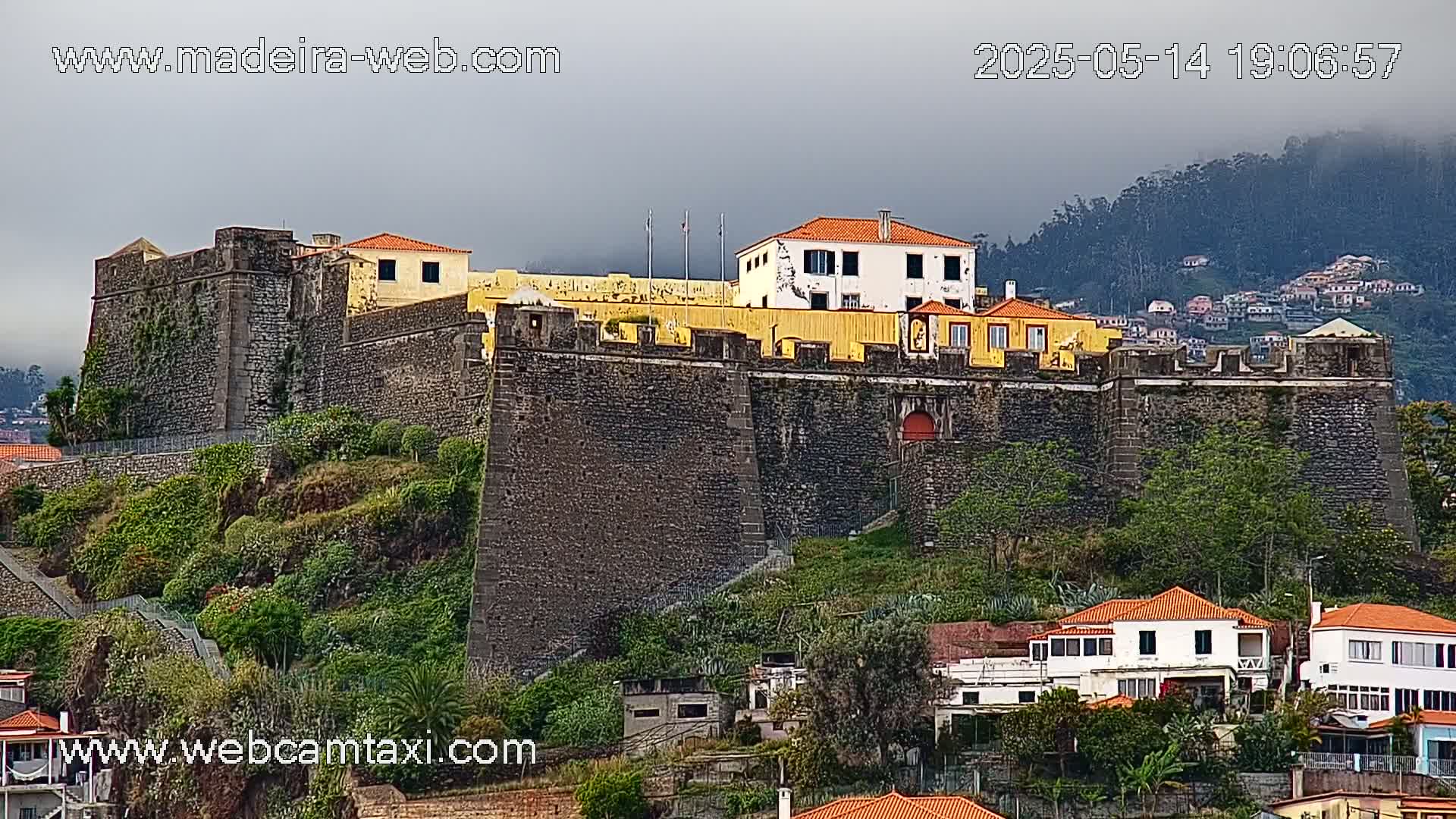 A large stone fortress with a yellow building sits atop a verdant hill under an overcast sky.