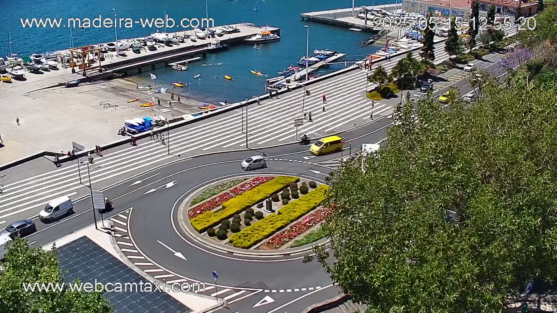 An aerial view shows a sunny day overlooking a harbor with boats, a paved walkway with pedestrians, and a roundabout with a colorful flower bed in the center, surrounded by cars and trees.