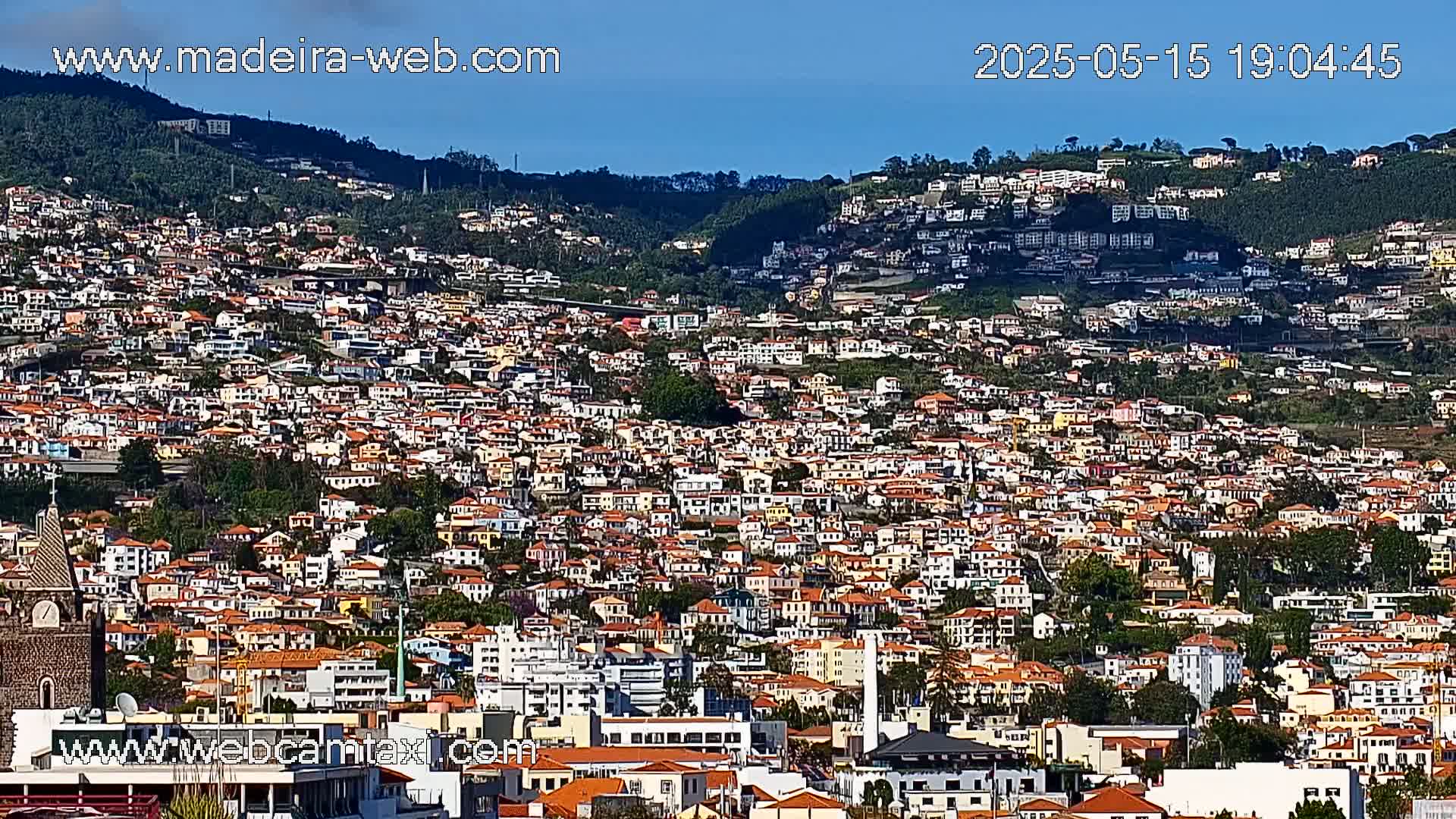 A densely populated hillside town with many low-rise buildings, mostly in shades of white, yellow, and orange, is visible under a clear blue sky.
