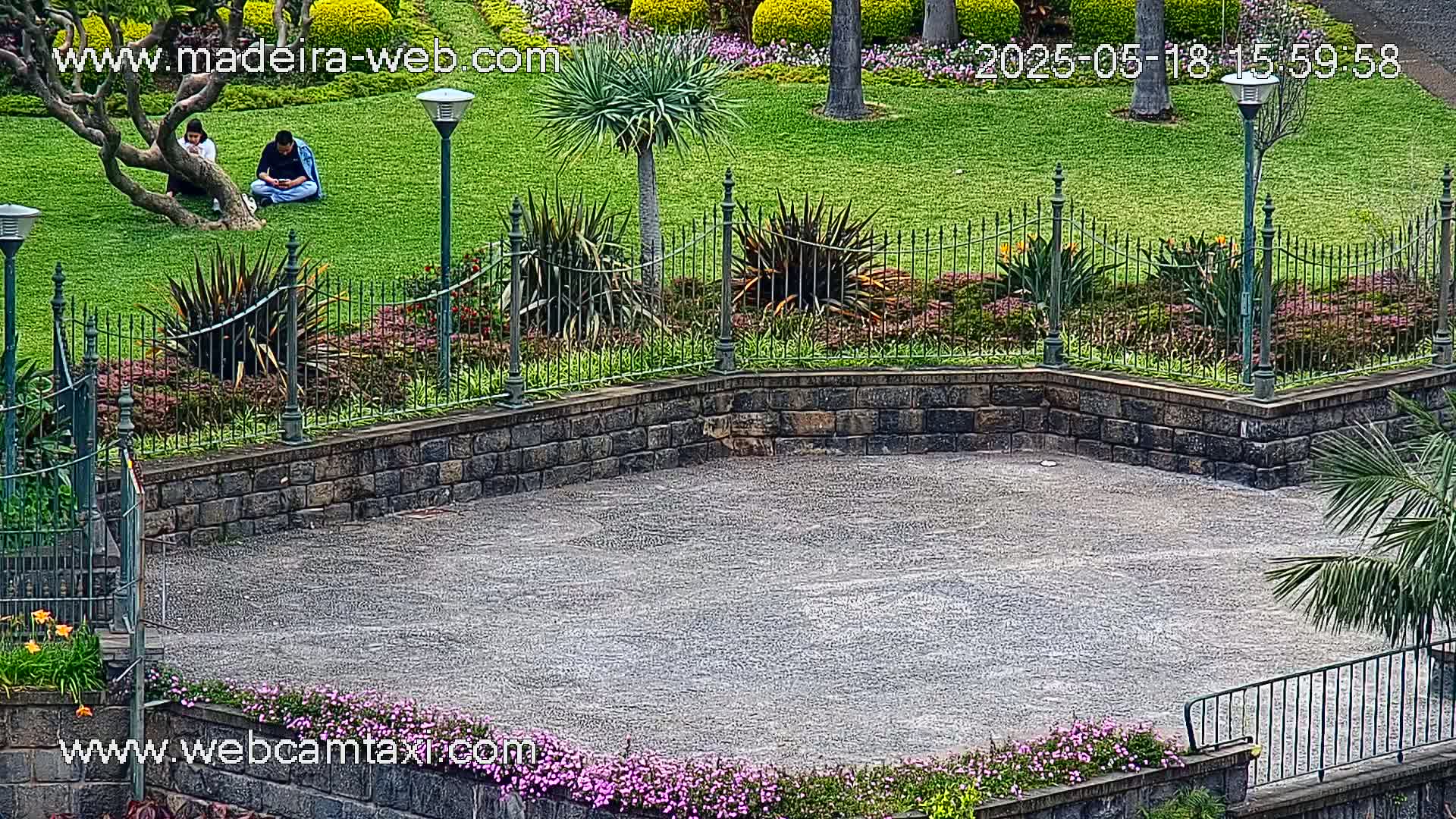 Two people sit on a grassy lawn surrounded by a stone wall, manicured gardens, and lampposts, on a cloudy day.