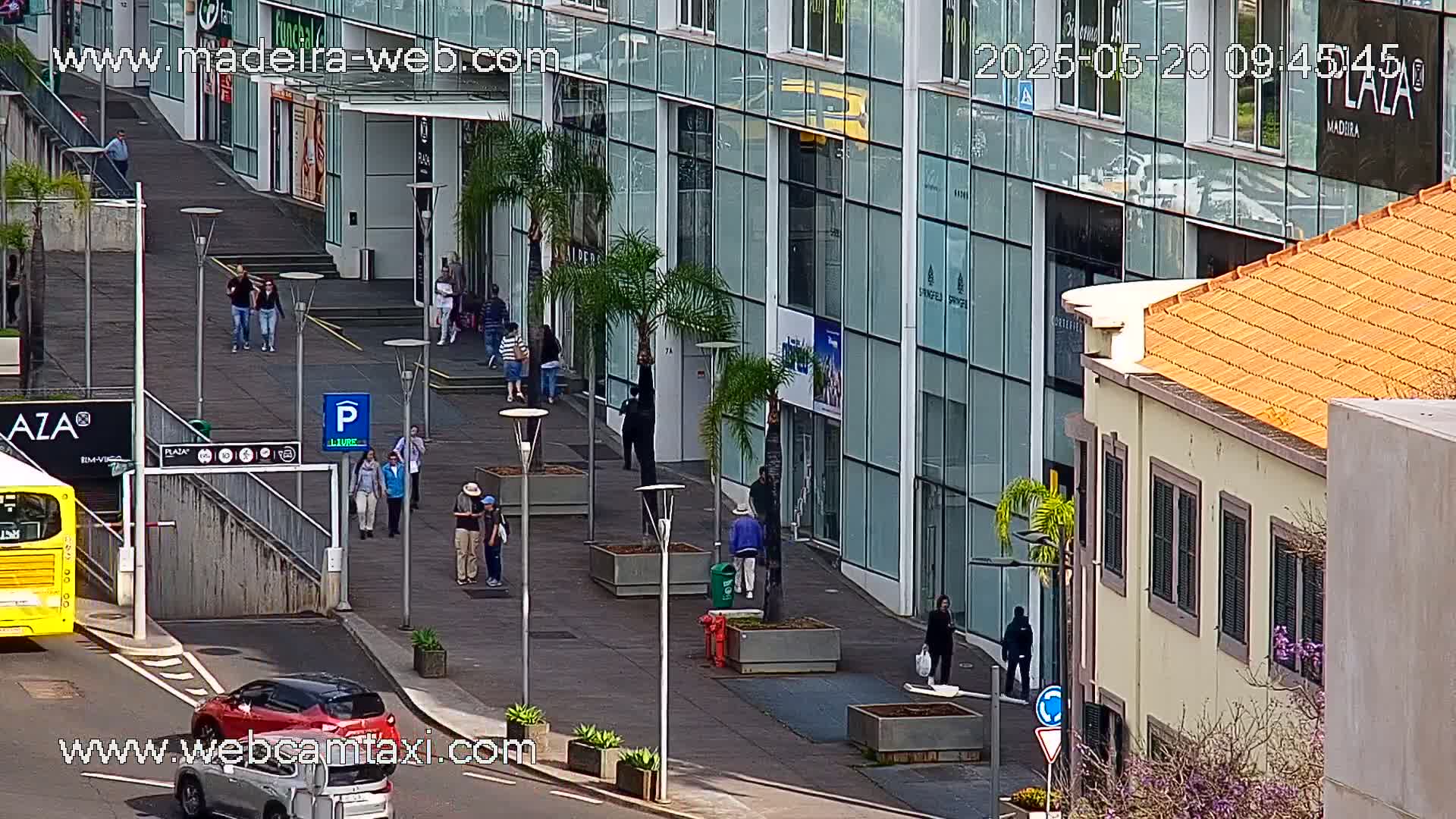 A city street scene with a modern glass building, pedestrians, and cars, under a sunny sky.