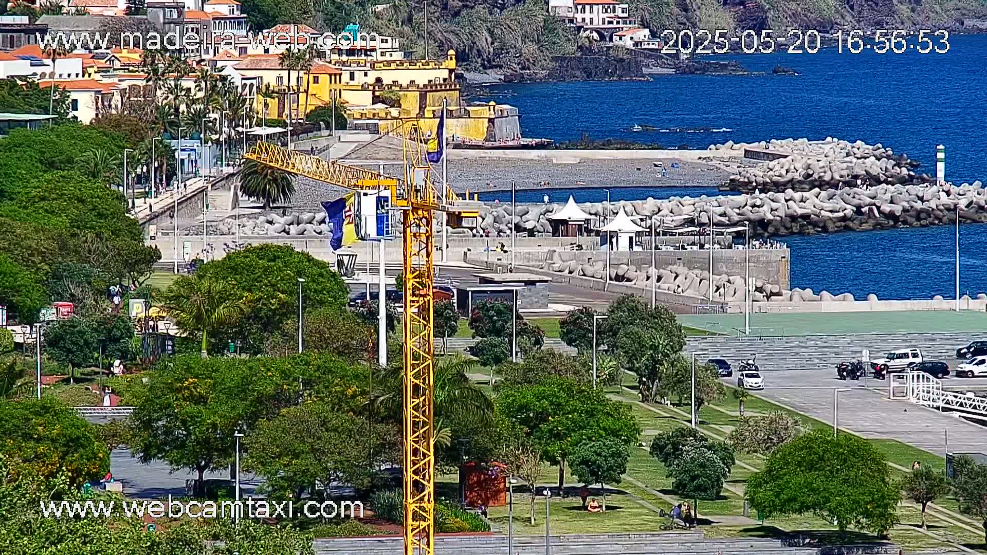 A coastal town scene on a sunny day shows a yellow crane next to a park with green lawns and trees, overlooking a rocky shoreline and calm blue ocean.