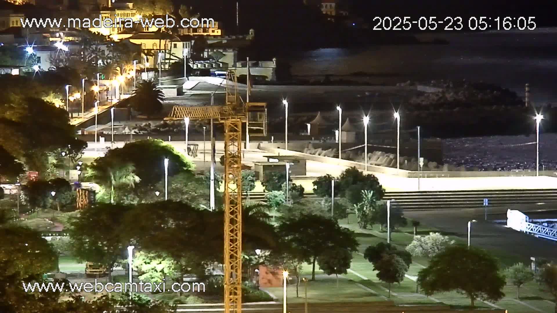 A nighttime view of a coastal park with a construction crane, illuminated by streetlights, overlooking a calm body of water.