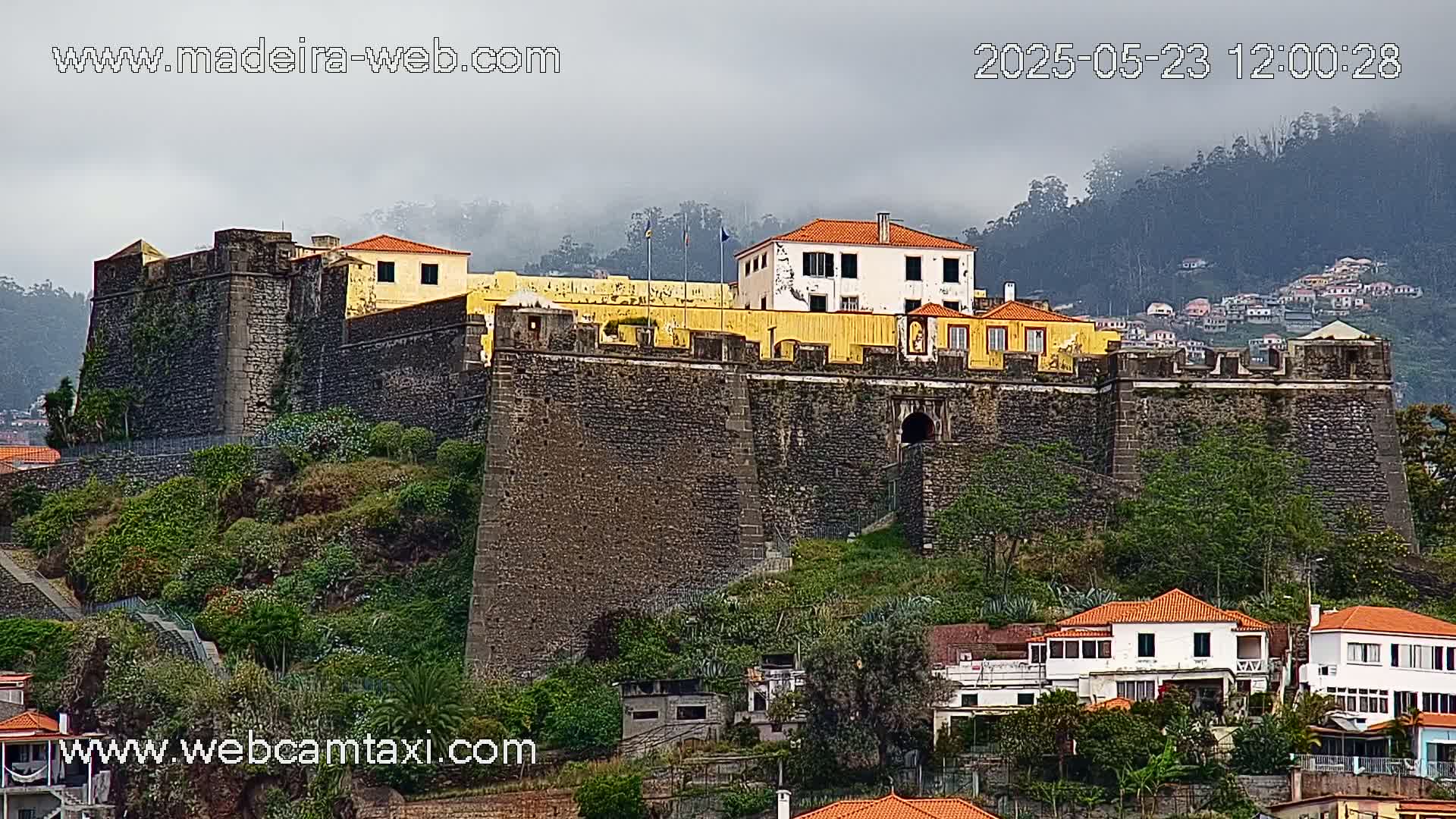 A large stone fortress with a yellow building sits atop a hill under an overcast sky, overlooking a town below.