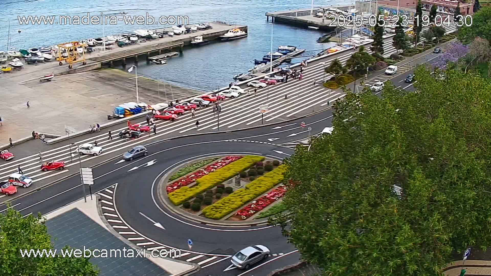 A roundabout with colorful landscaping is surrounded by roads with several cars, some parked and others driving, near a harbor with boats and calm water under a partly cloudy sky.