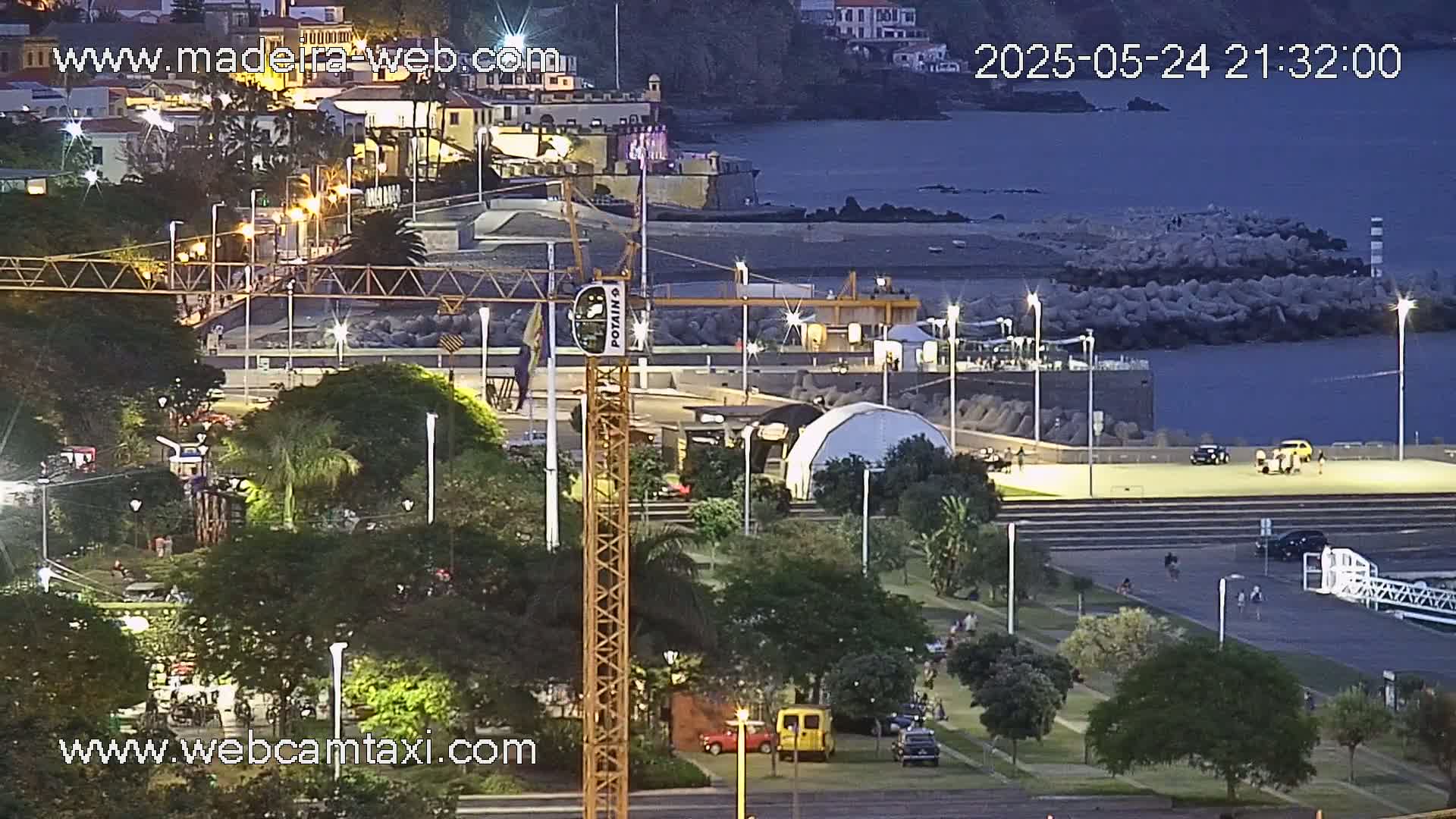 A nighttime view of a coastal city park with a construction crane, vehicles, and people, overlooking a calm ocean under a dark twilight sky.