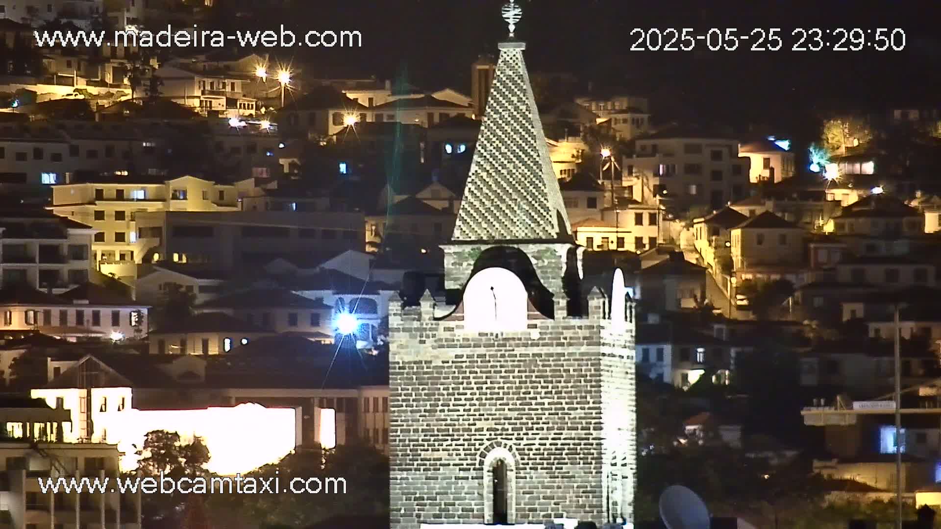 A stone tower stands prominently against a backdrop of a hillside town illuminated at night.