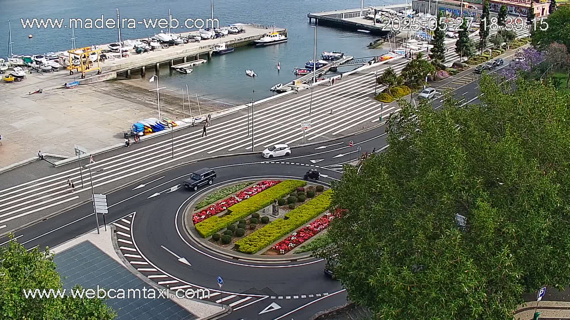 An aerial view shows a harbor with boats, a roundabout with colorful landscaping, and several cars driving on roads with pedestrian crosswalks, on a mostly sunny day.