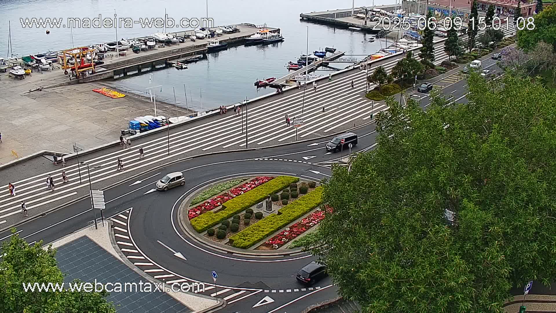 An aerial view shows a harbor with boats, a pedestrian crossing with people walking, a roundabout with a landscaped center, and cars driving on roads, all under an overcast sky.