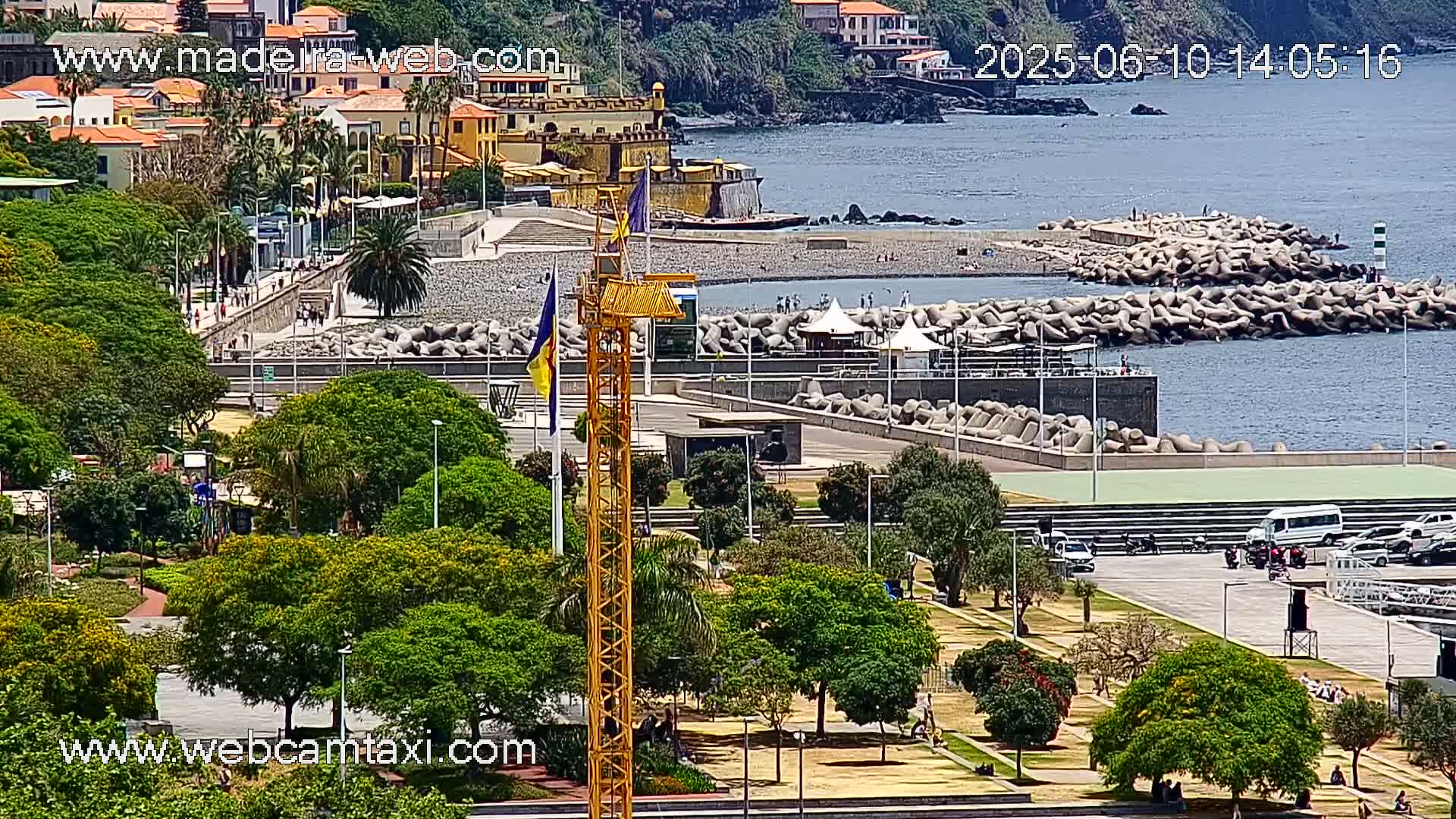 A coastal scene on a sunny day shows a park with lush green trees, a paved area with parked vehicles, a breakwater protecting a pebble beach, and buildings along the hillside in the background.