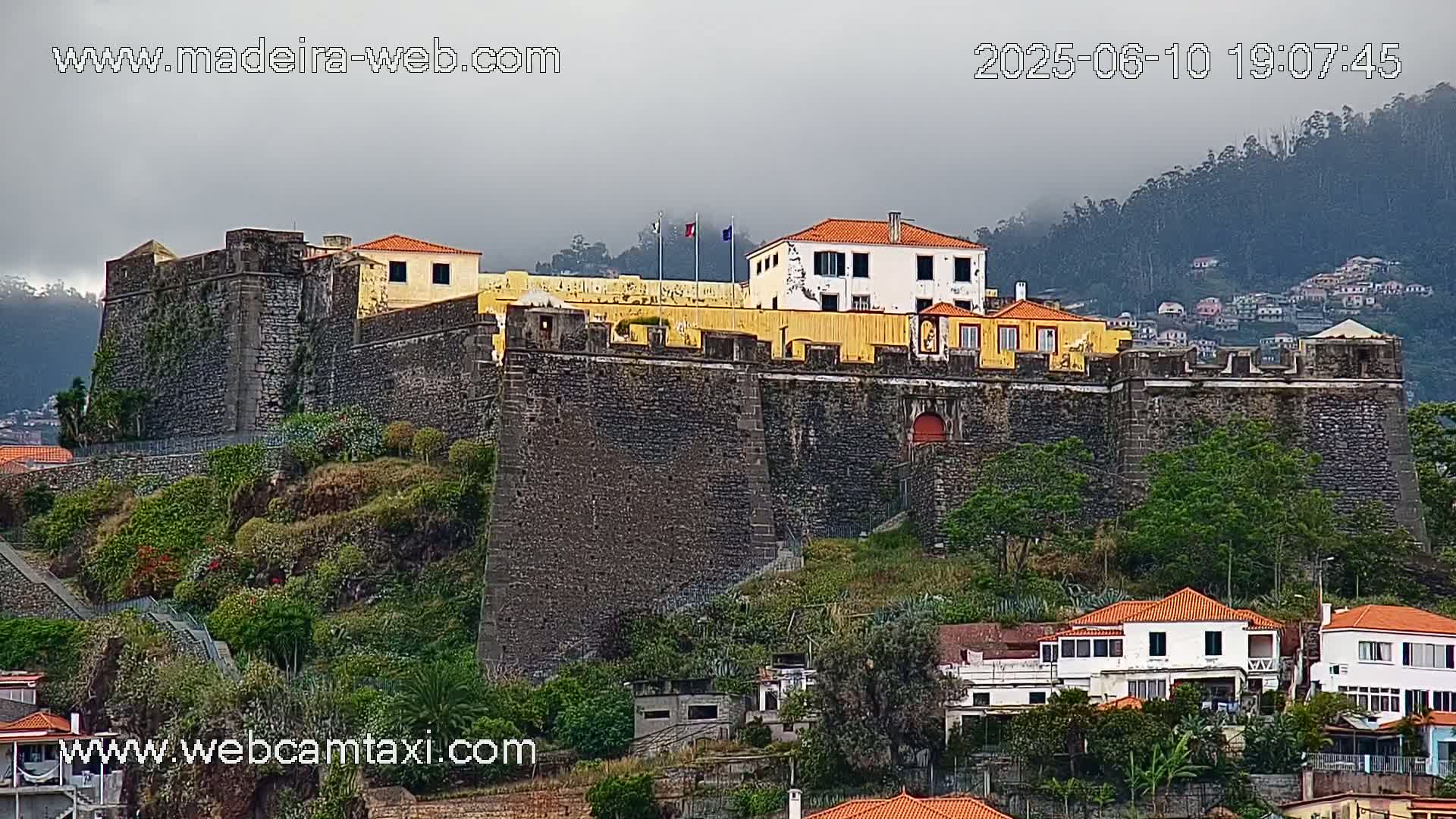 A stone fortress with a yellow building at its center sits atop a hill on an overcast day, overlooking a village of white and orange-roofed houses.