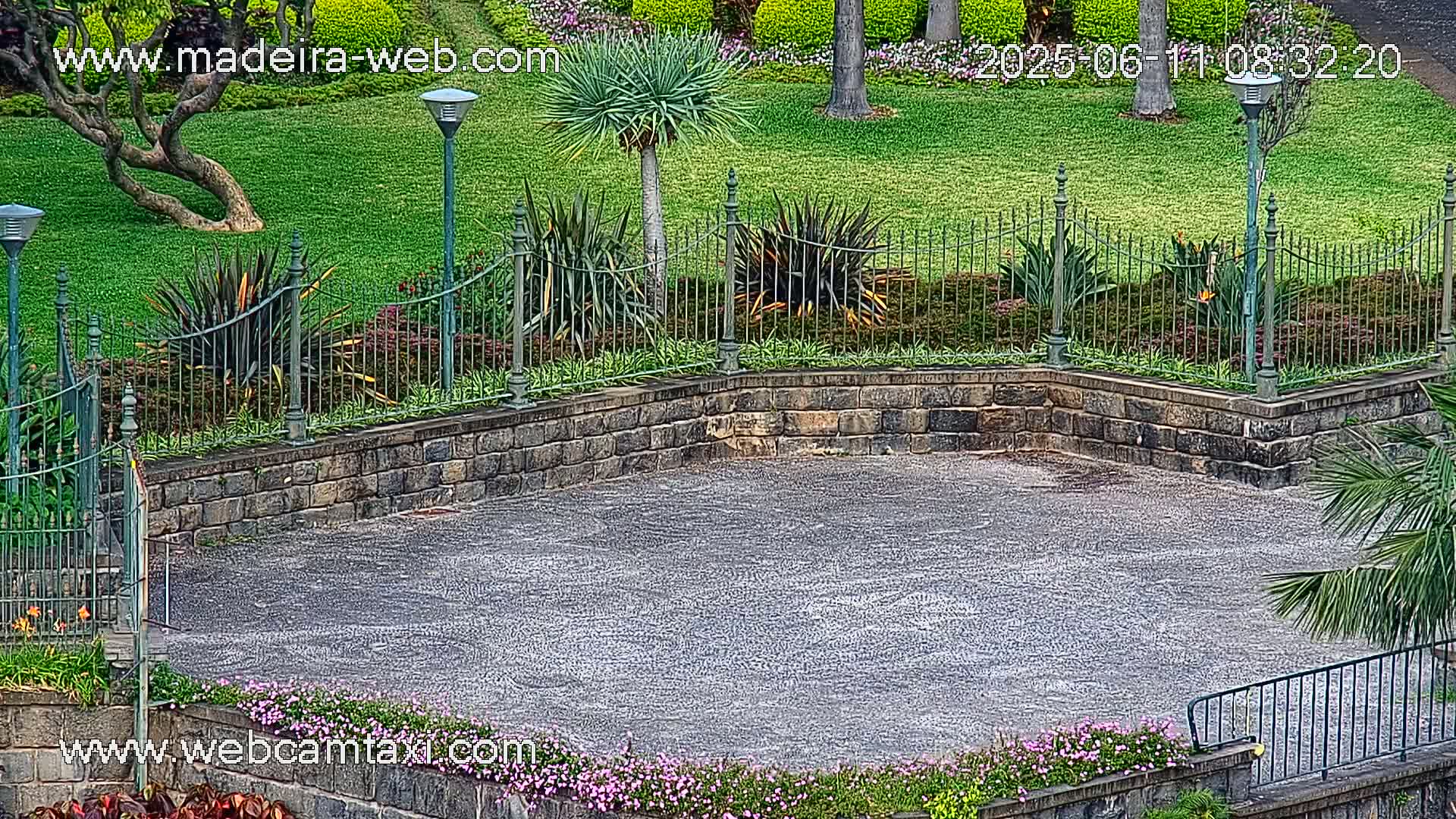 A paved area bordered by a stone wall and wrought-iron fence, surrounded by lush green lawns and tropical plants under an overcast sky.