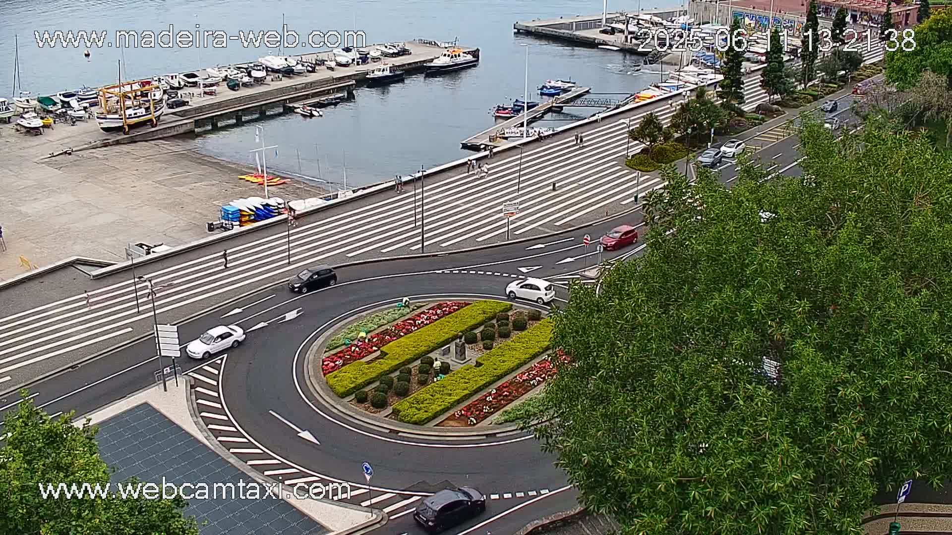 A harbor with boats is visible in the distance, beyond a roundabout with a landscaped center, surrounded by a pedestrian area with crosswalks and several cars driving on the road.
