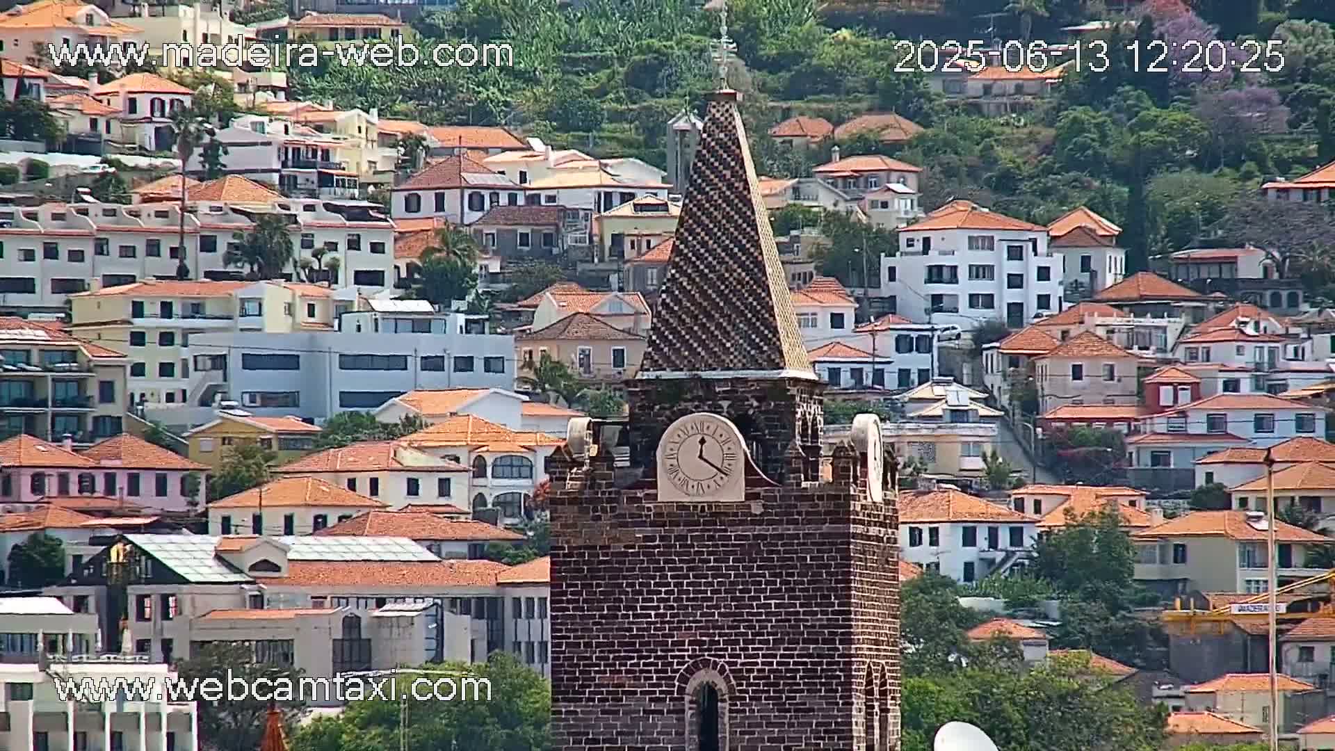 A brick clock tower with a patterned spire stands prominently in front of a densely packed hillside of red-roofed buildings on a sunny day.