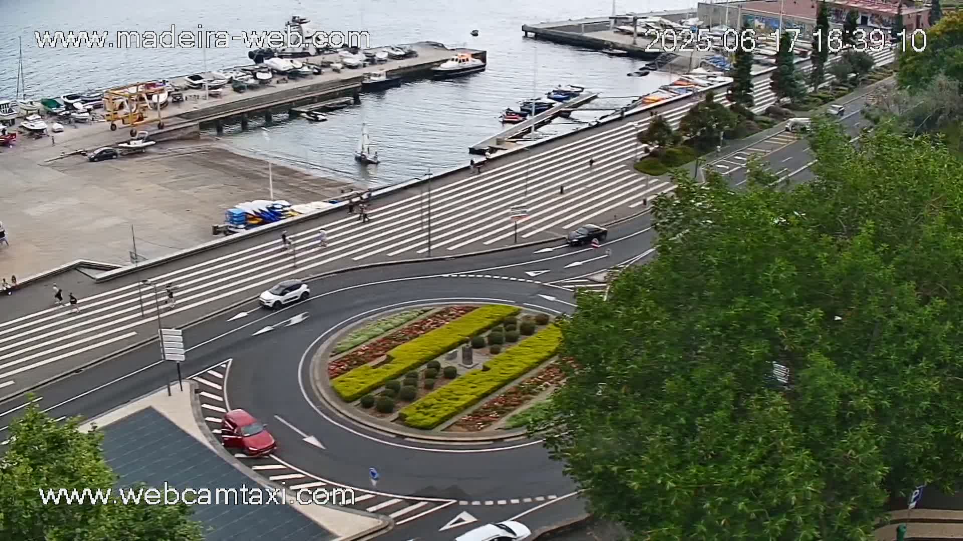An aerial view shows a harbor with boats, a pedestrian walkway with crosswalks, a roundabout with a landscaped center, and several cars on the road, on a partly sunny day.