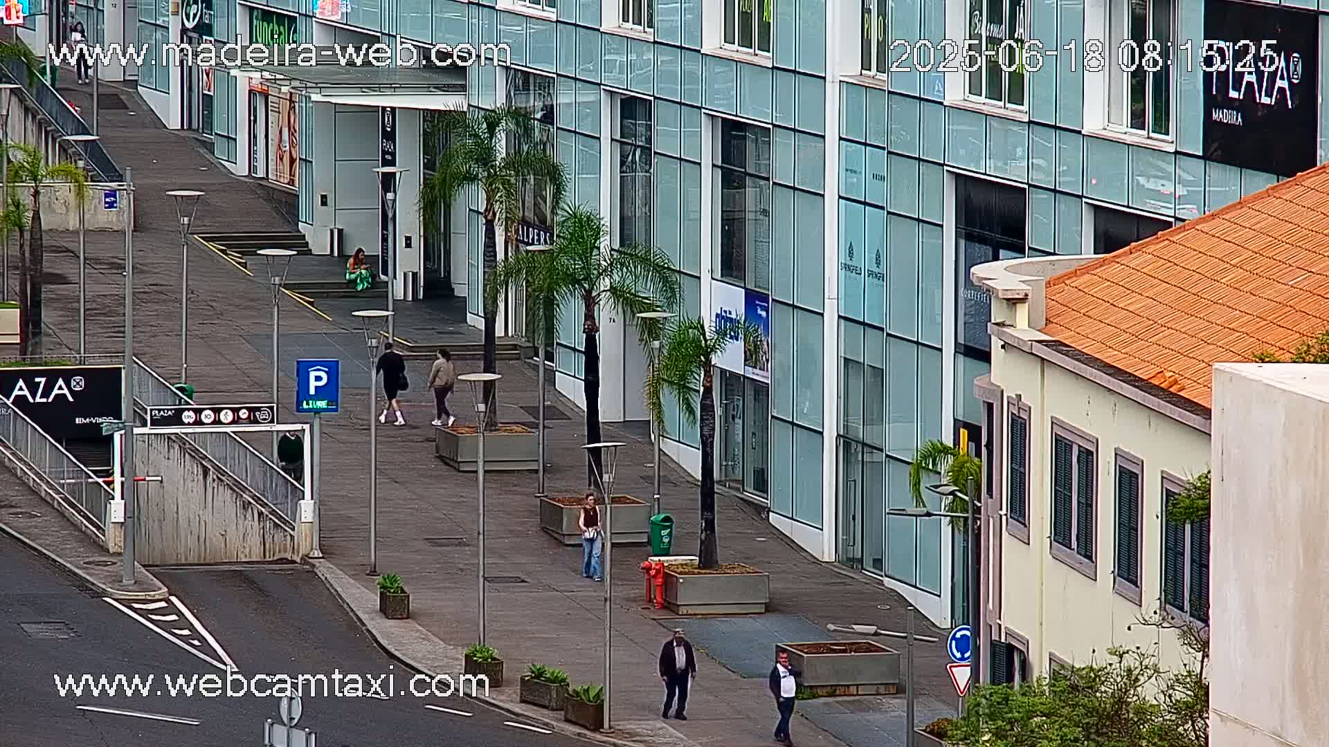 A paved pedestrian area in front of modern glass buildings and a smaller, older building, with a few people walking around on an overcast day.