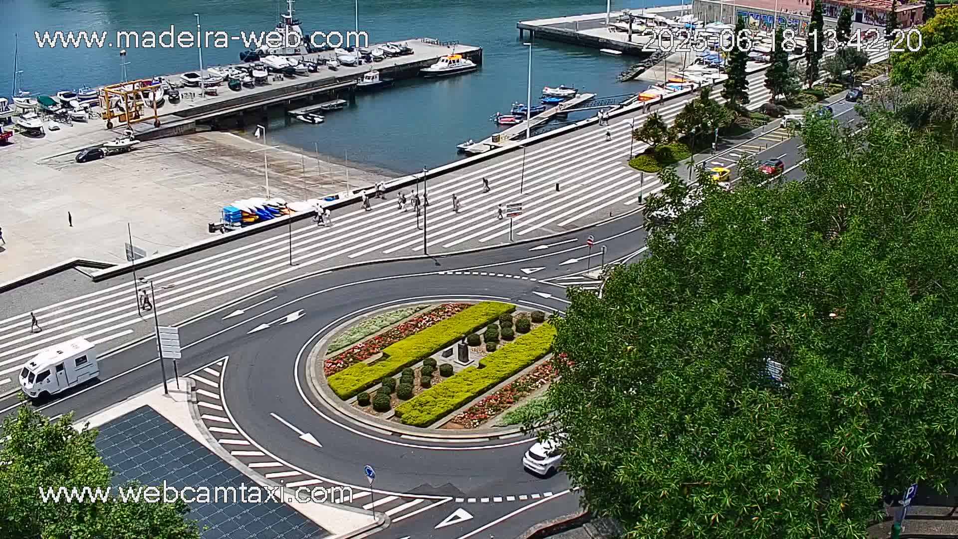 A sunny day overlooking a harbor with boats, a roundabout with a landscaped center, and a roadway with pedestrians and vehicles.