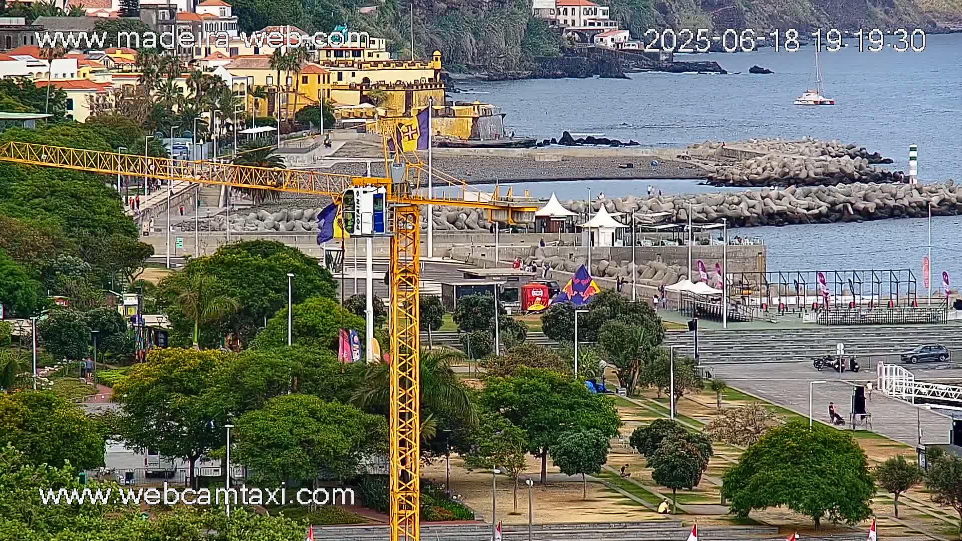 A coastal town scene with a yellow crane, a park, a breakwater, a calm ocean, and buildings on a hillside under mostly sunny skies.