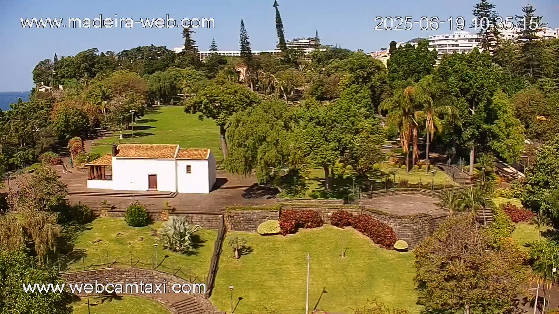 A sunny aerial view of a park with a small white building, lush greenery, and palm trees, situated near buildings and ocean in the distance.