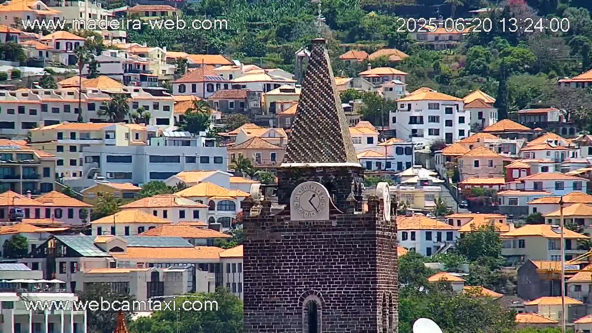 A brick clock tower with a tiled spire stands prominently in the foreground, overlooking a densely packed hillside village of low-rise buildings with orange tile roofs on a sunny day.
