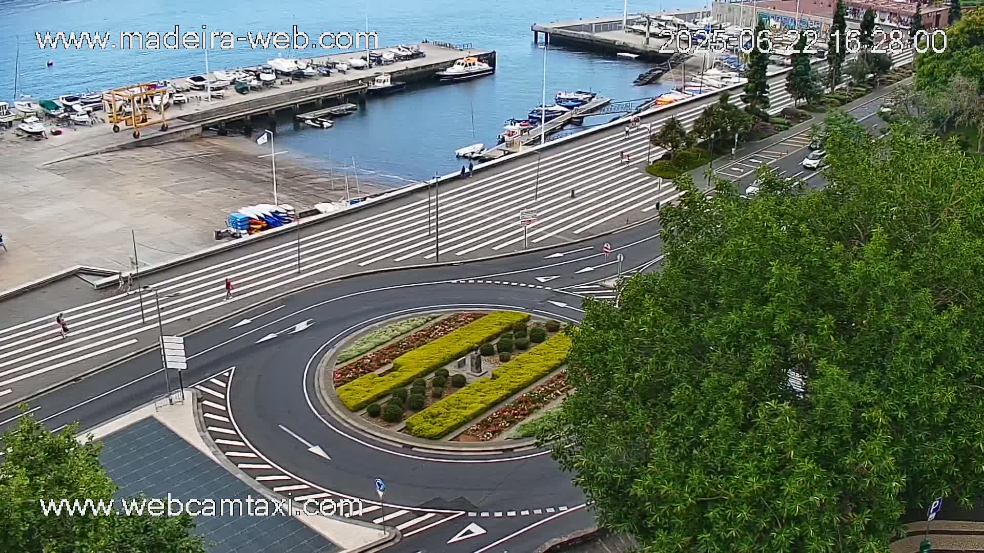 An aerial view shows a harbor with boats, a roadway with a roundabout featuring landscaped center, and a calm body of water under a partly cloudy sky.
