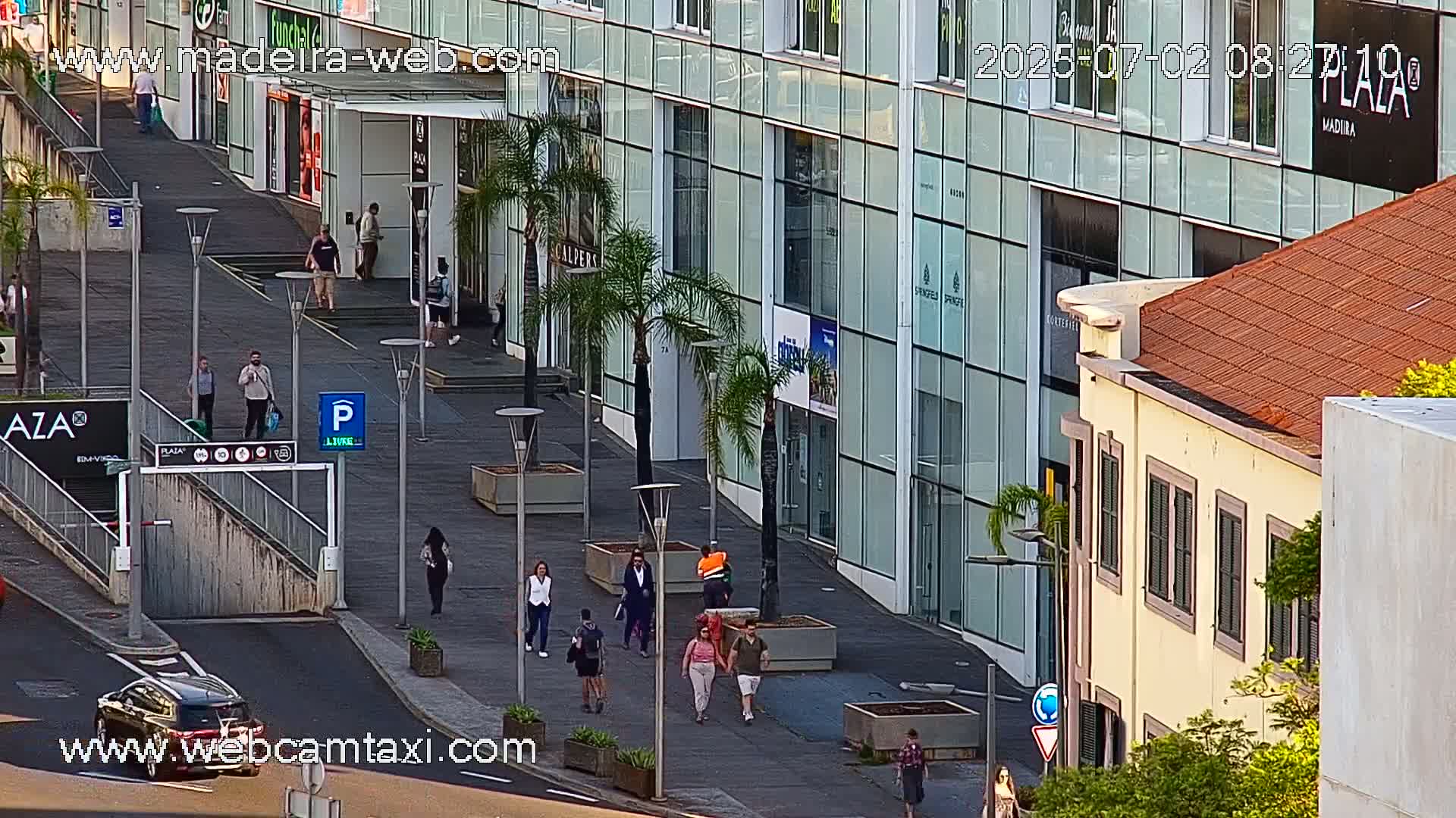 A mostly sunny outdoor scene shows pedestrians walking on a paved walkway in front of modern glass buildings and a smaller, older building with a terracotta roof; a car is driving on the street.