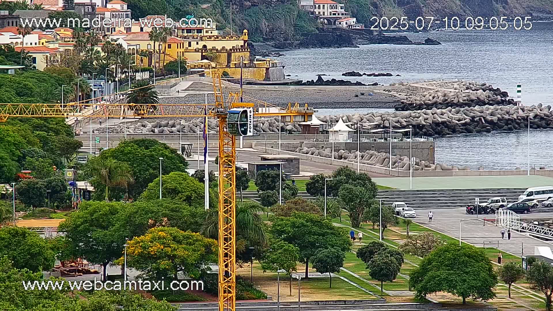 A yellow crane stands in a park overlooking a coastline with a breakwater, buildings, and a calm ocean under an overcast sky.