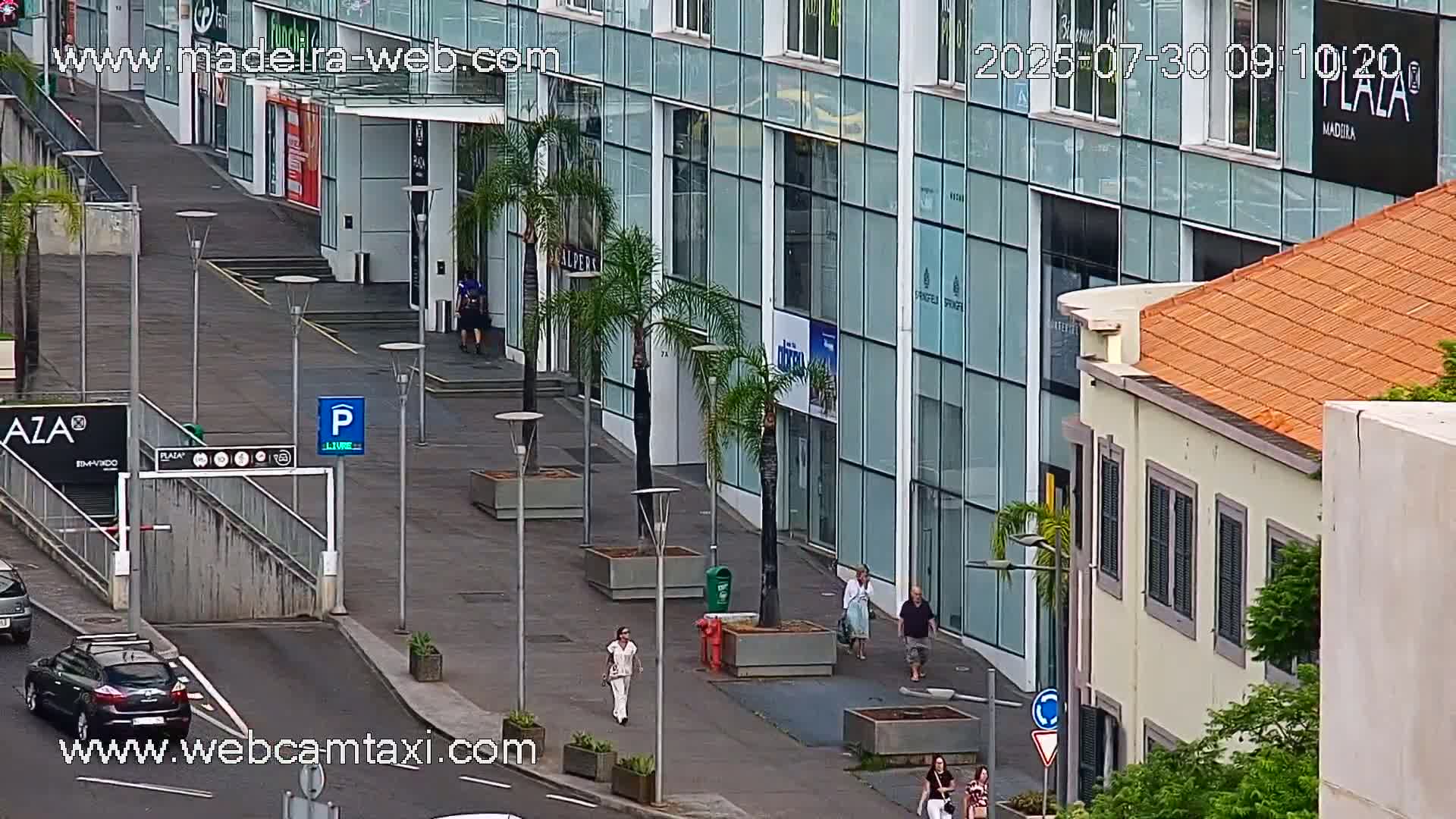 A city street scene on a partly sunny day shows pedestrians walking past shops and palm trees, with cars parked and driving along the roadway.