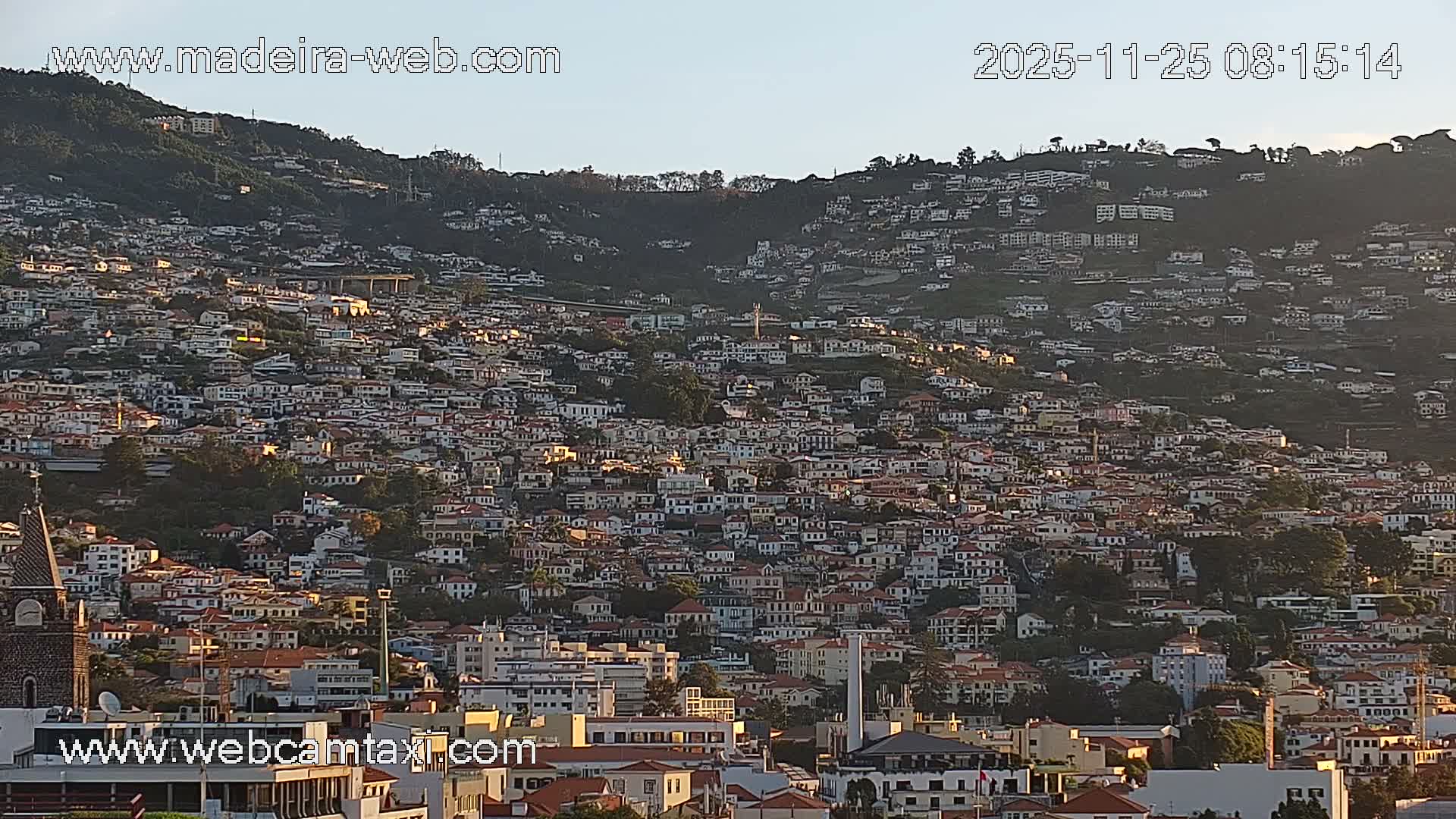 A panoramic view reveals a densely built city of white and terracotta-roofed houses sprawling across a verdant mountainside, bathed in bright sunlight under a clear, blue sky.