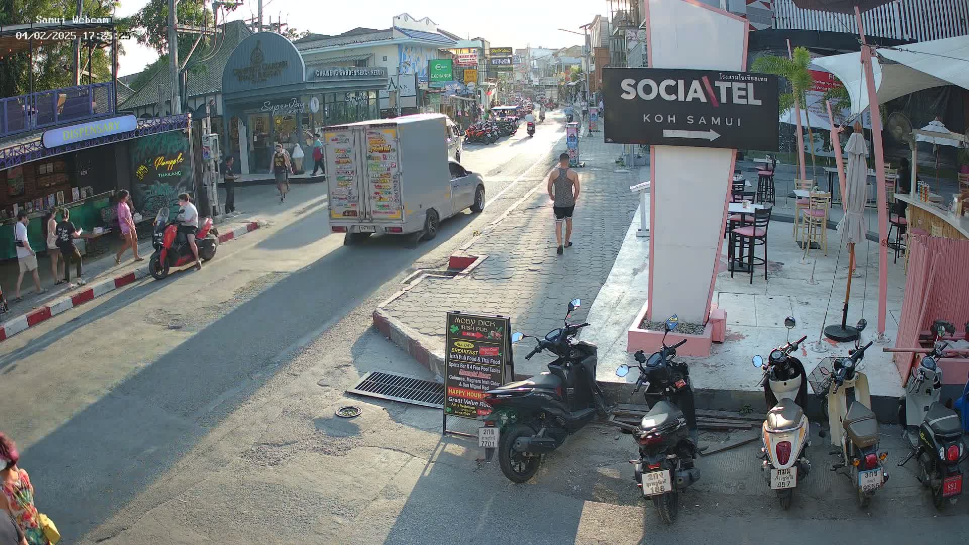 A sunny street scene shows pedestrians walking and several motorbikes parked along the roadside, with a truck and other vehicles driving on the road.