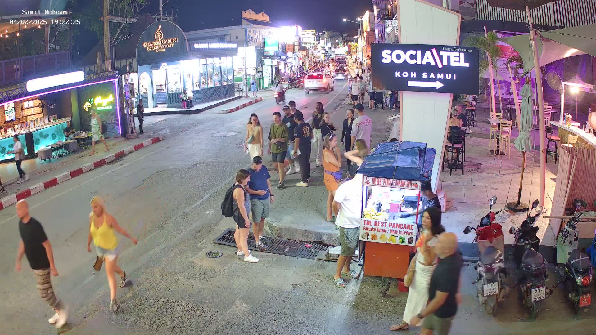 A nighttime street scene shows numerous people walking along a street lined with shops and restaurants, with a food vendor visible in the foreground and several parked motorbikes.