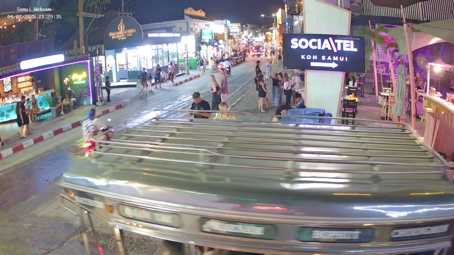 A nighttime street scene shows a bustling pedestrian area with shops and restaurants, viewed from a moving vehicle, under clear skies.