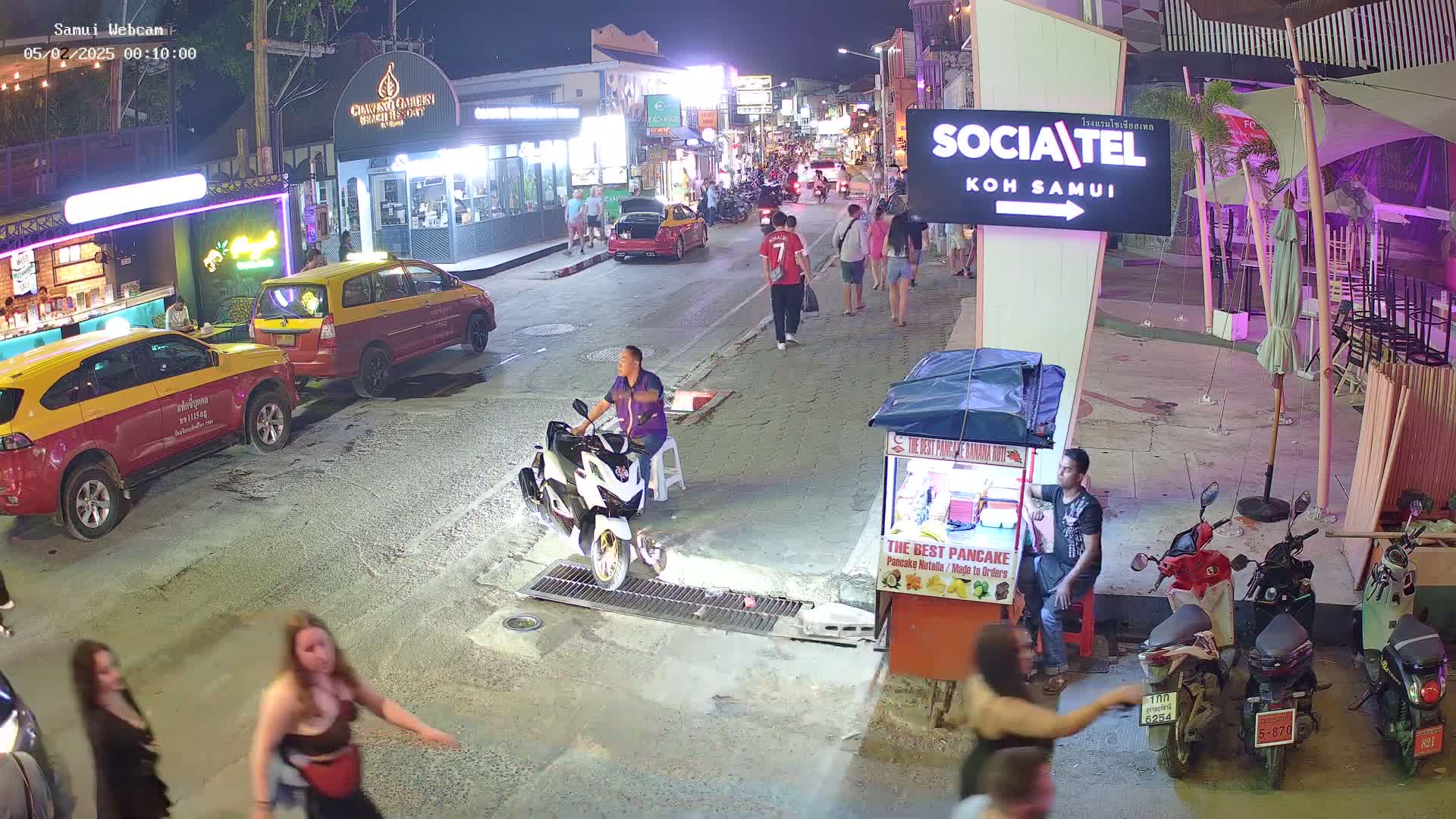 A nighttime street scene shows pedestrians and vehicles, including taxis and motorbikes, near food stalls and businesses under artificial street lights.