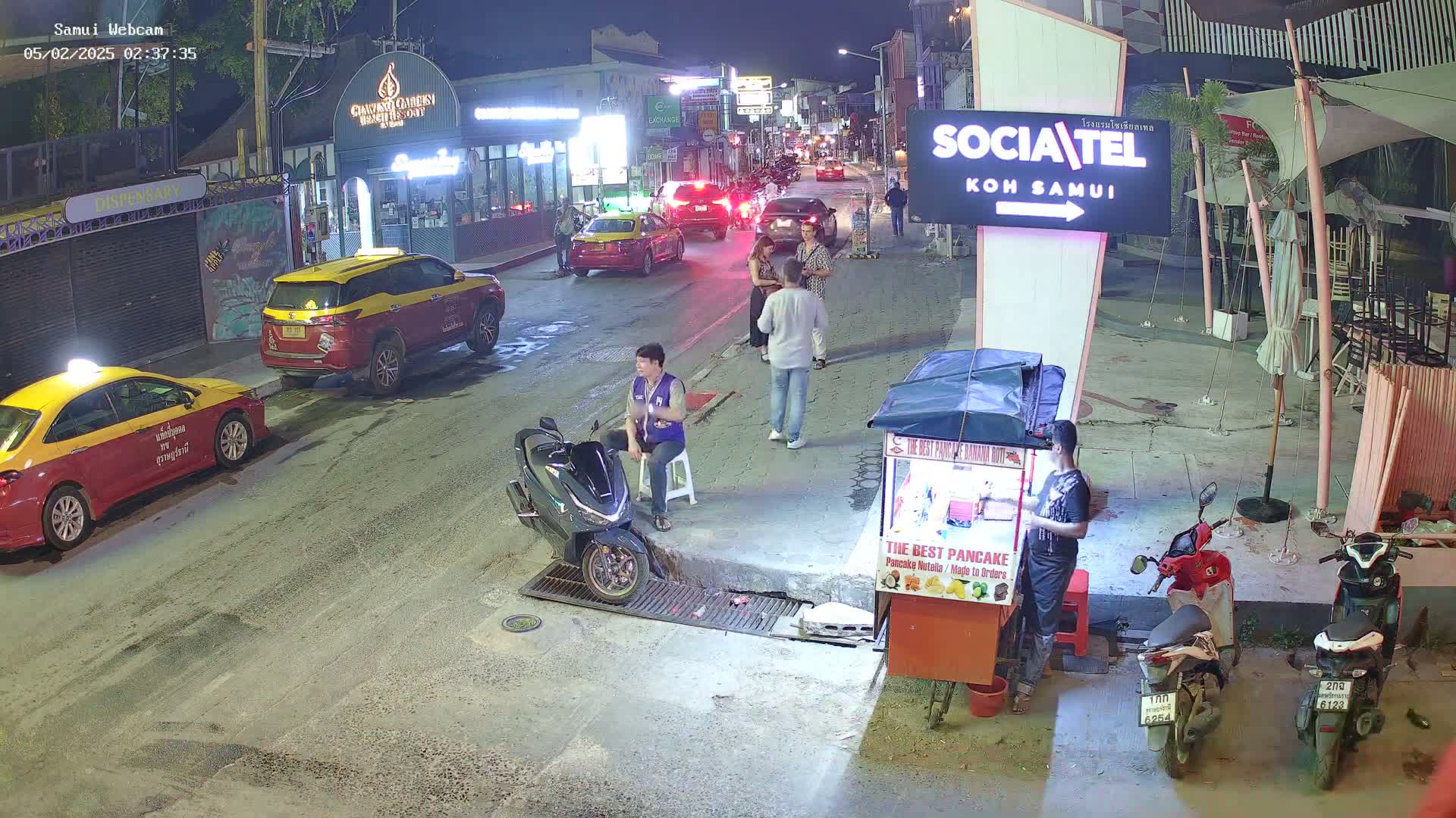 A nighttime street scene shows several people standing around parked cars and scooters, with a food vendor cart nearby.