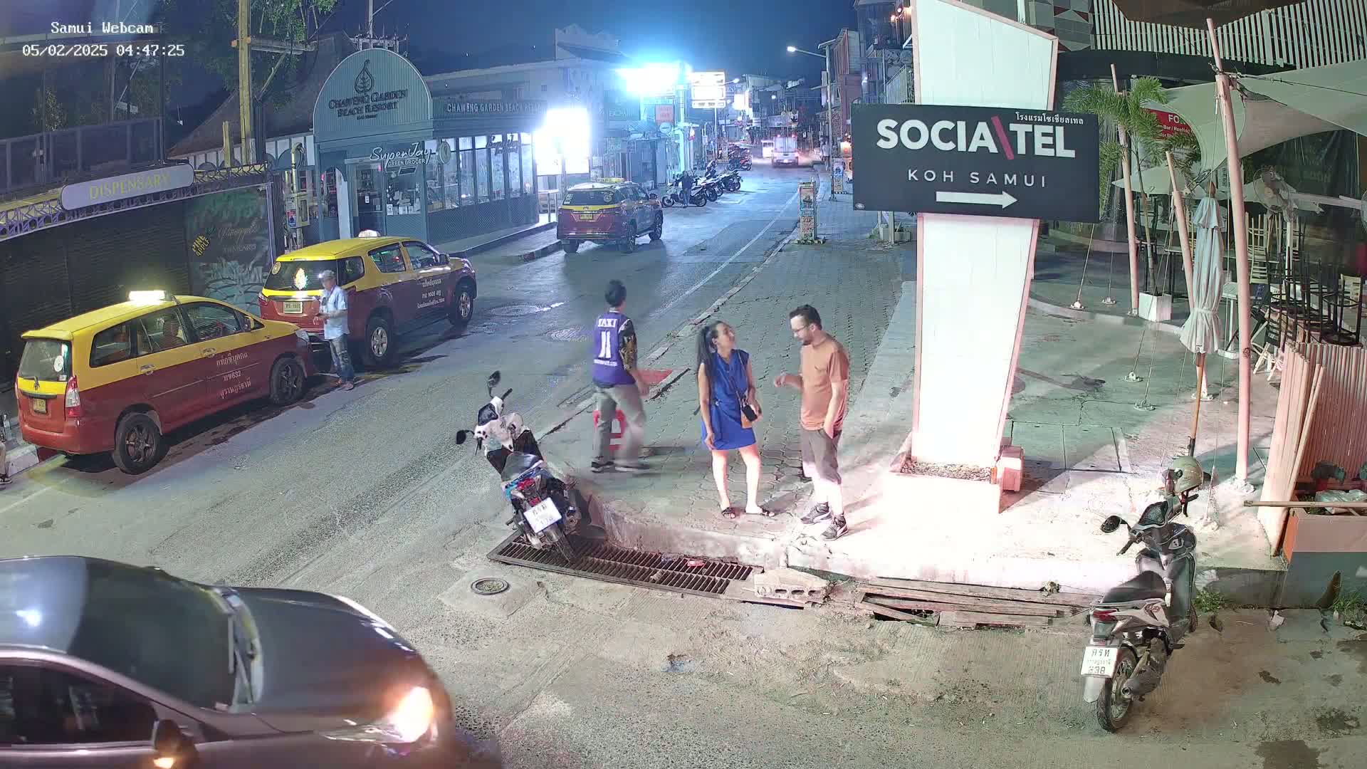 A nighttime street scene shows several people standing and talking near parked taxis and motorbikes, under the light of street lamps.