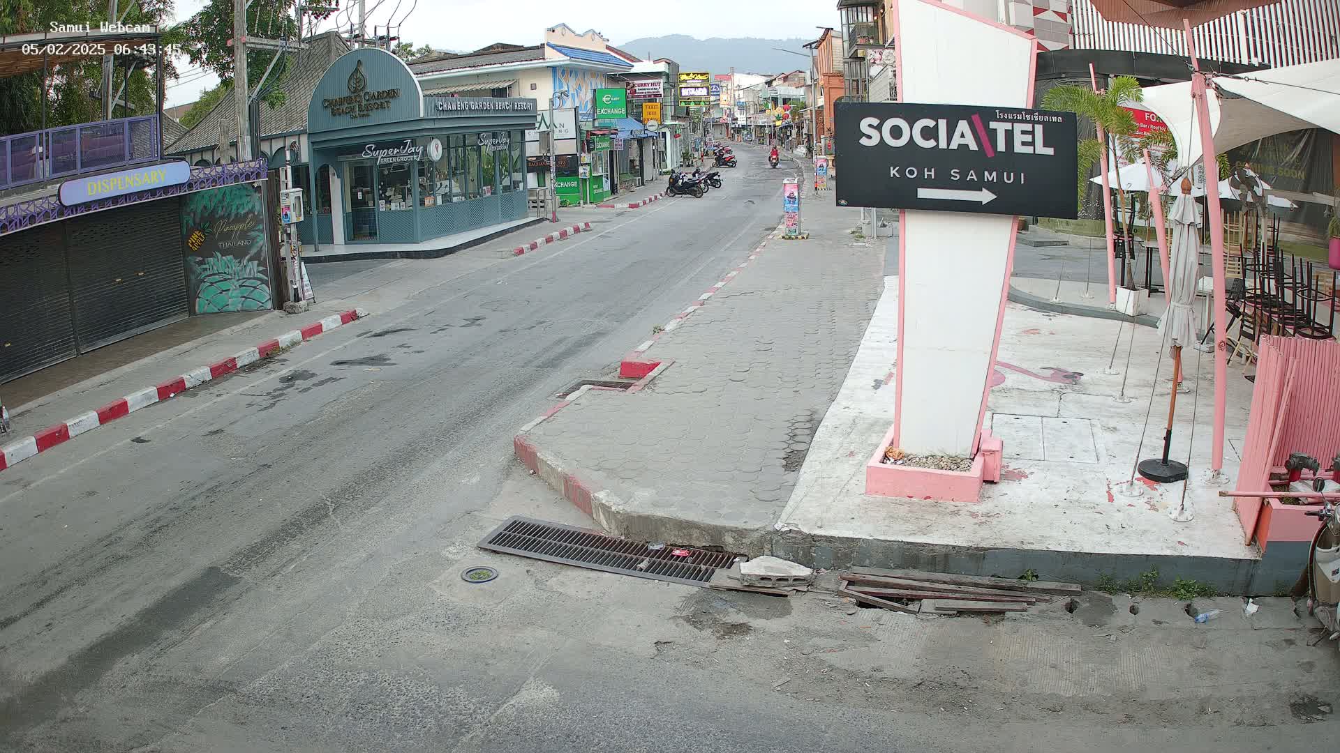A mostly empty street scene in Koh Samui shows several shops and restaurants, some with outdoor seating, under an overcast sky.