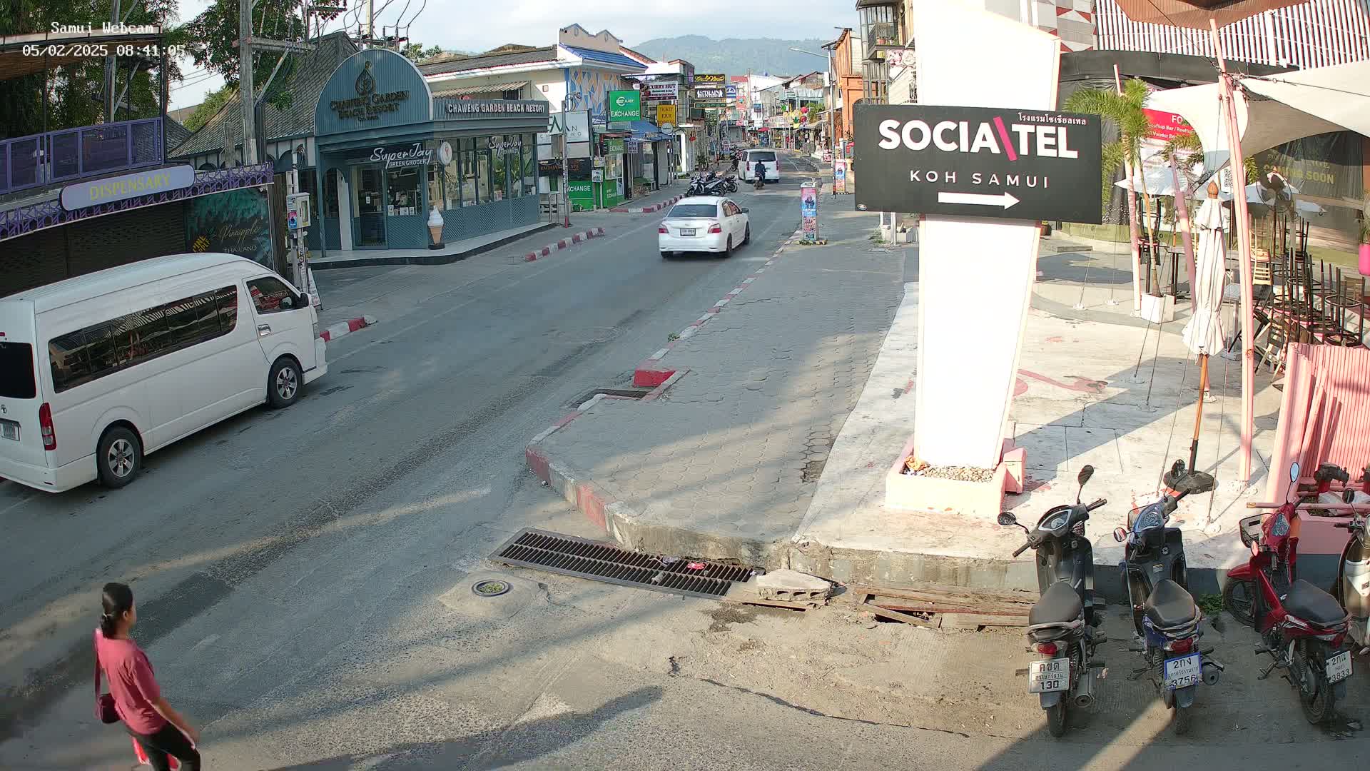 A sunny street scene shows a white van and car parked on the side of a road, with several motorcycles parked near a restaurant and a pedestrian walking.