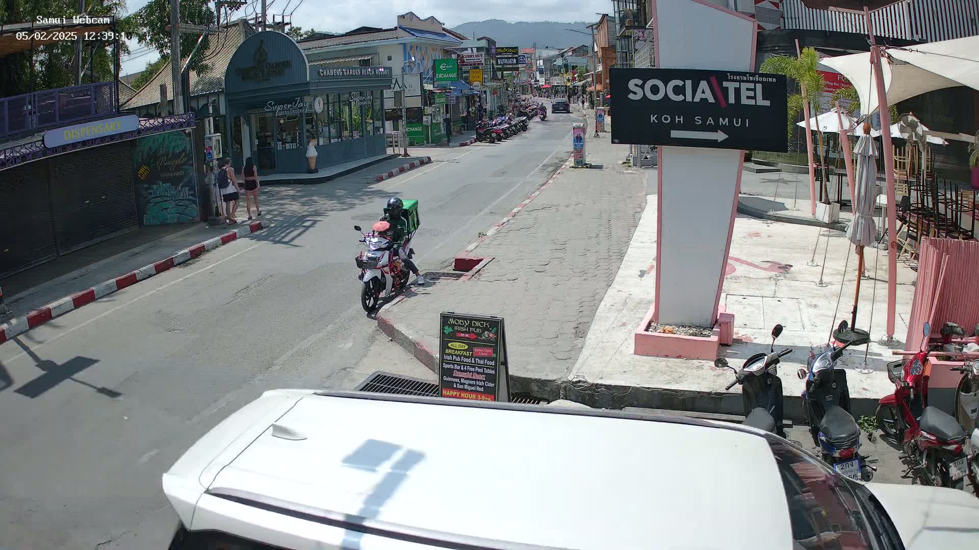 A sunny street scene in a town shows pedestrians, parked motorbikes, and a delivery person on a motorbike; several shops line the street.