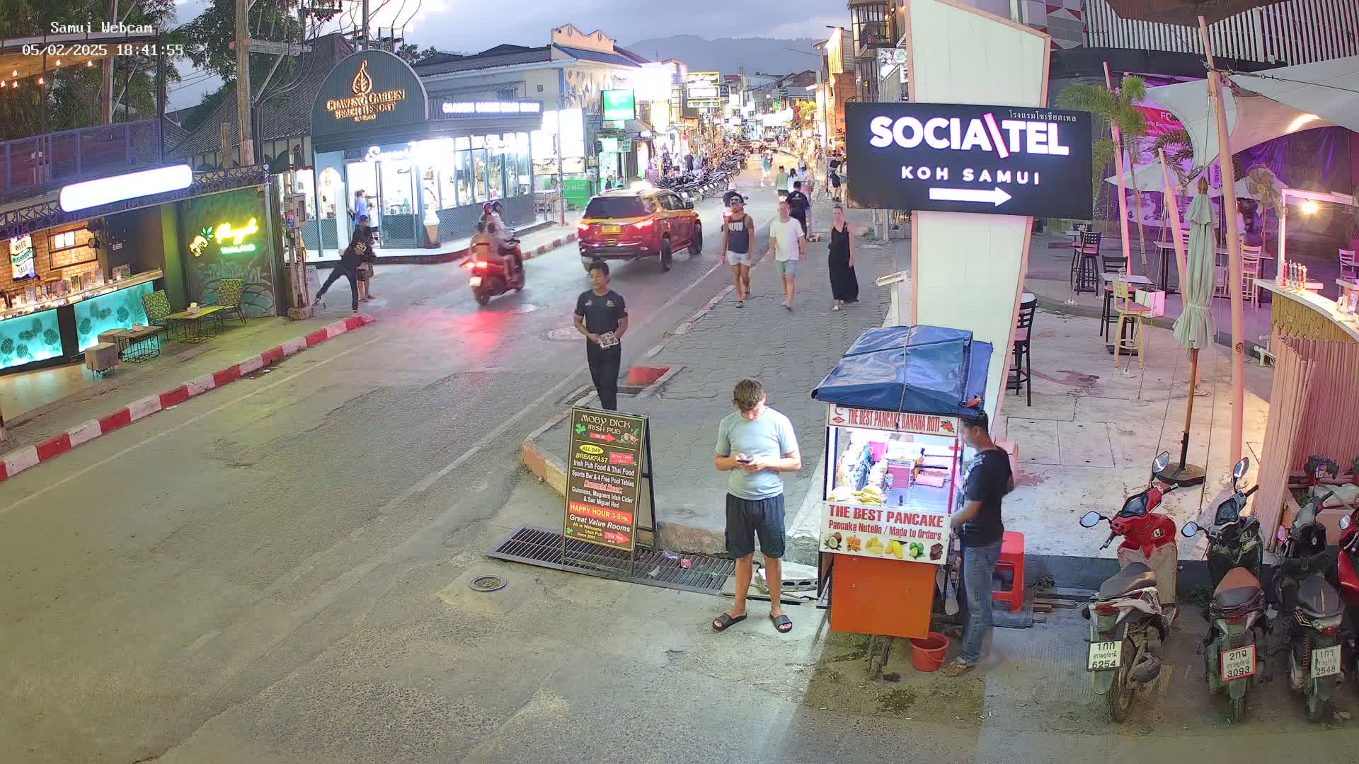 A street scene at dusk shows people walking past shops and restaurants, with several parked motorbikes and a food stand.