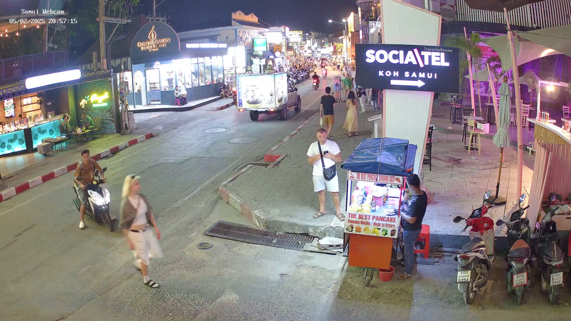 A nighttime street scene shows people walking and riding motorbikes past shops and restaurants, with a food cart and parked motorbikes nearby.