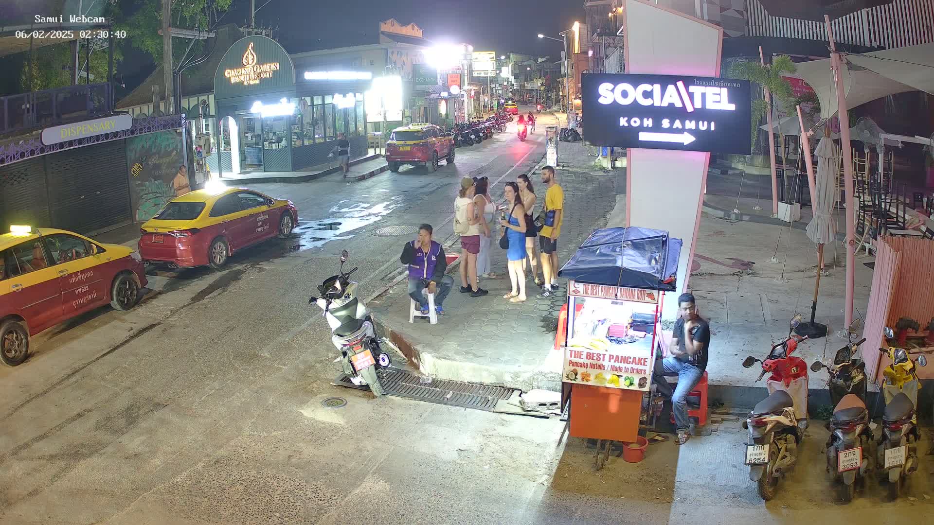 At night, several people stand in a street with parked taxis and motorbikes near food stalls and businesses under bright lights.