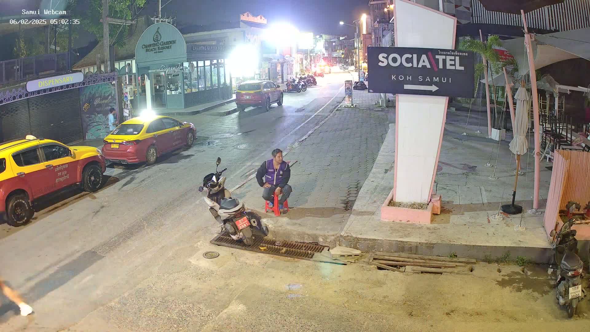 A man sits on a red stool next to a parked scooter on a nighttime street in front of a restaurant, with two yellow taxis parked nearby and other vehicles visible down the street.