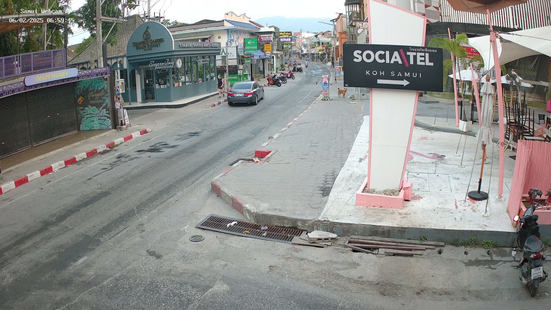 A street scene on a partly cloudy day shows a road with cars and motorcycles, pedestrians, and businesses along the sides, including a dispensary and a restaurant with outdoor seating.