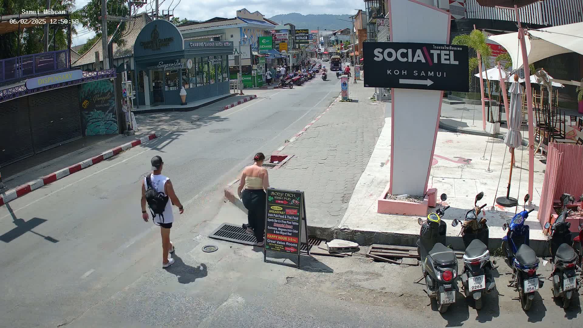 A sunny street scene shows two pedestrians walking on a paved road lined with shops and parked motorbikes, under a mostly clear sky.